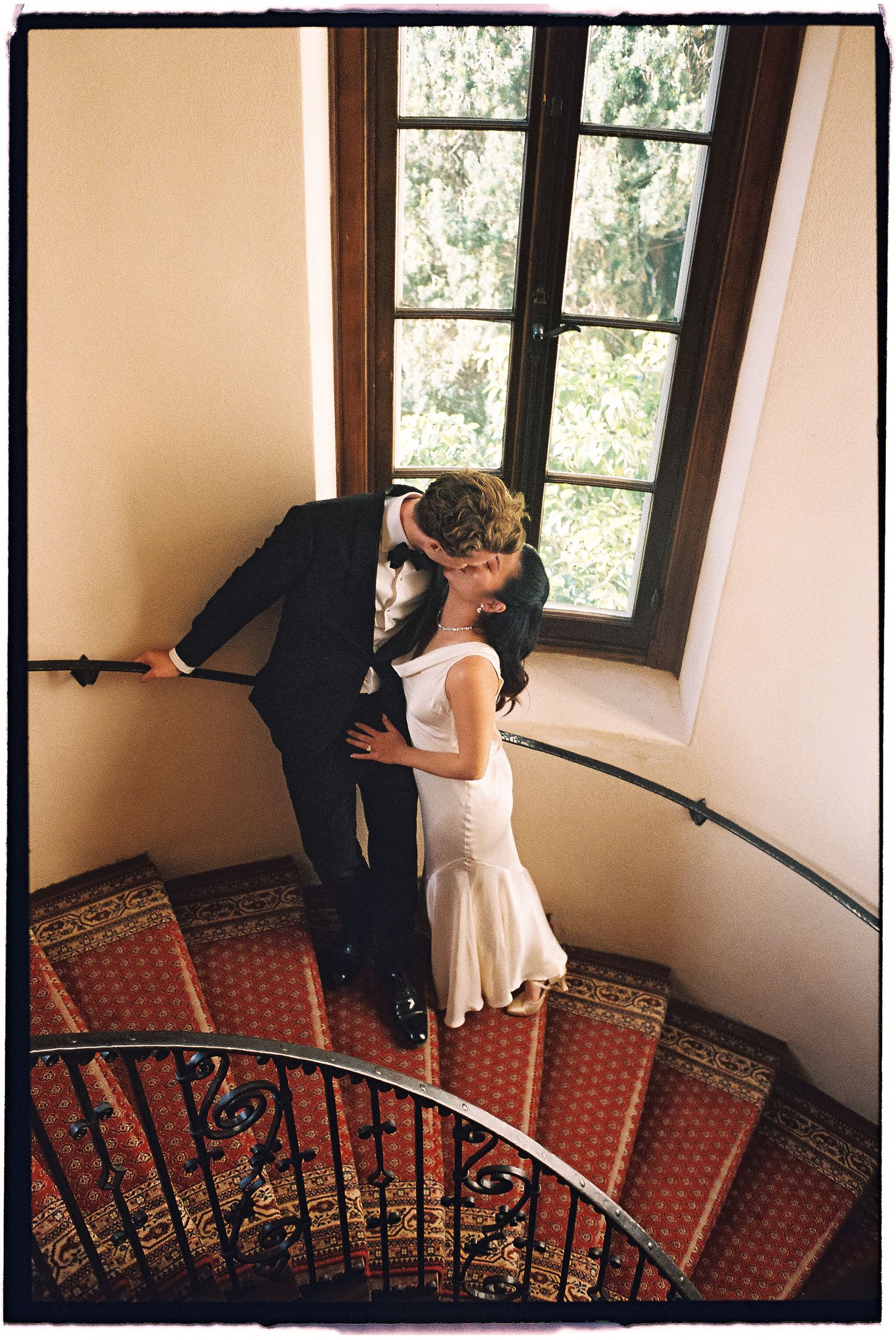 Bride and groom sharing a romantic kiss on an elegant curved staircase with red carpet runner near a large window.