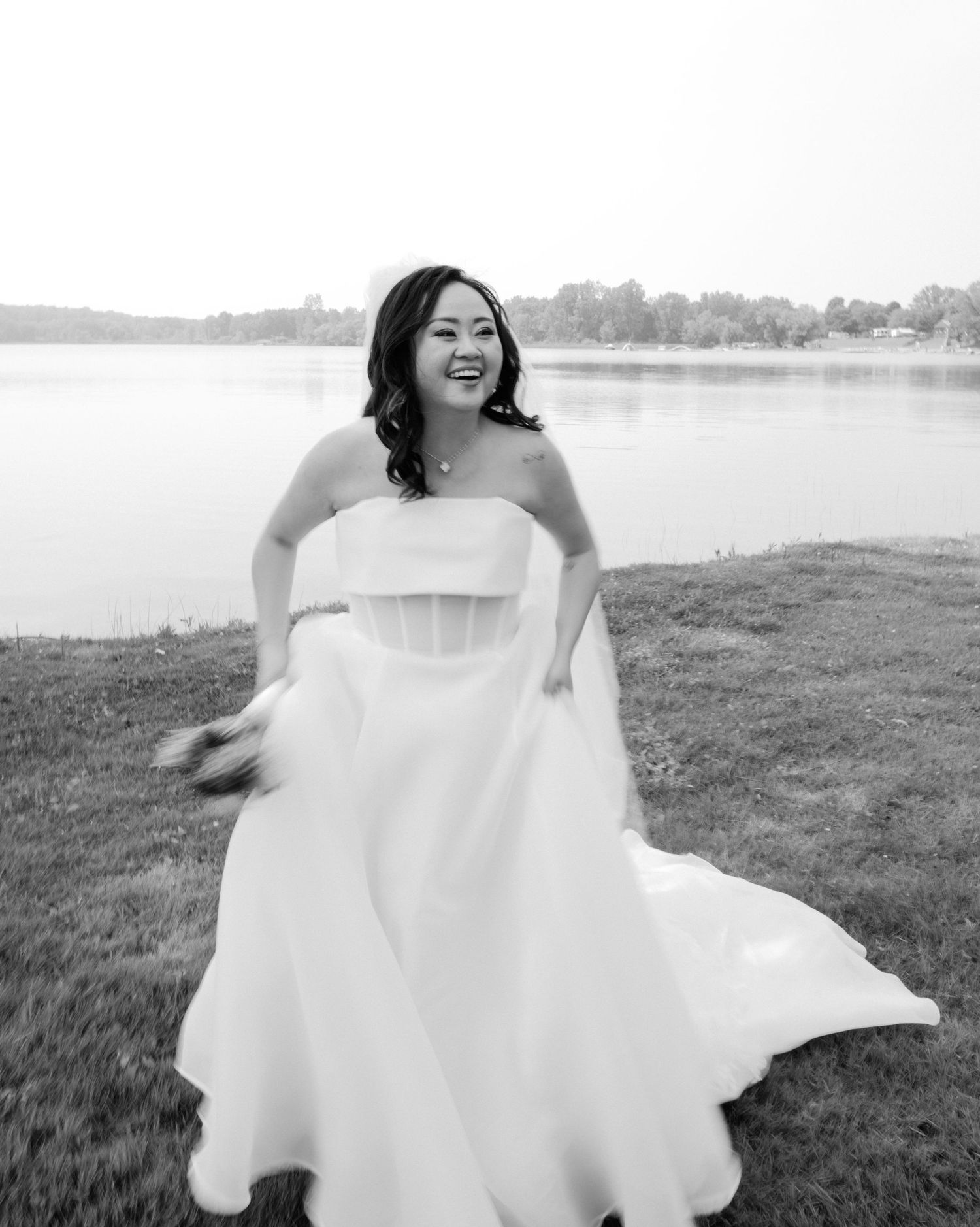 A joyful bride in a white strapless wedding dress twirls on grass near a serene lake in black and white.