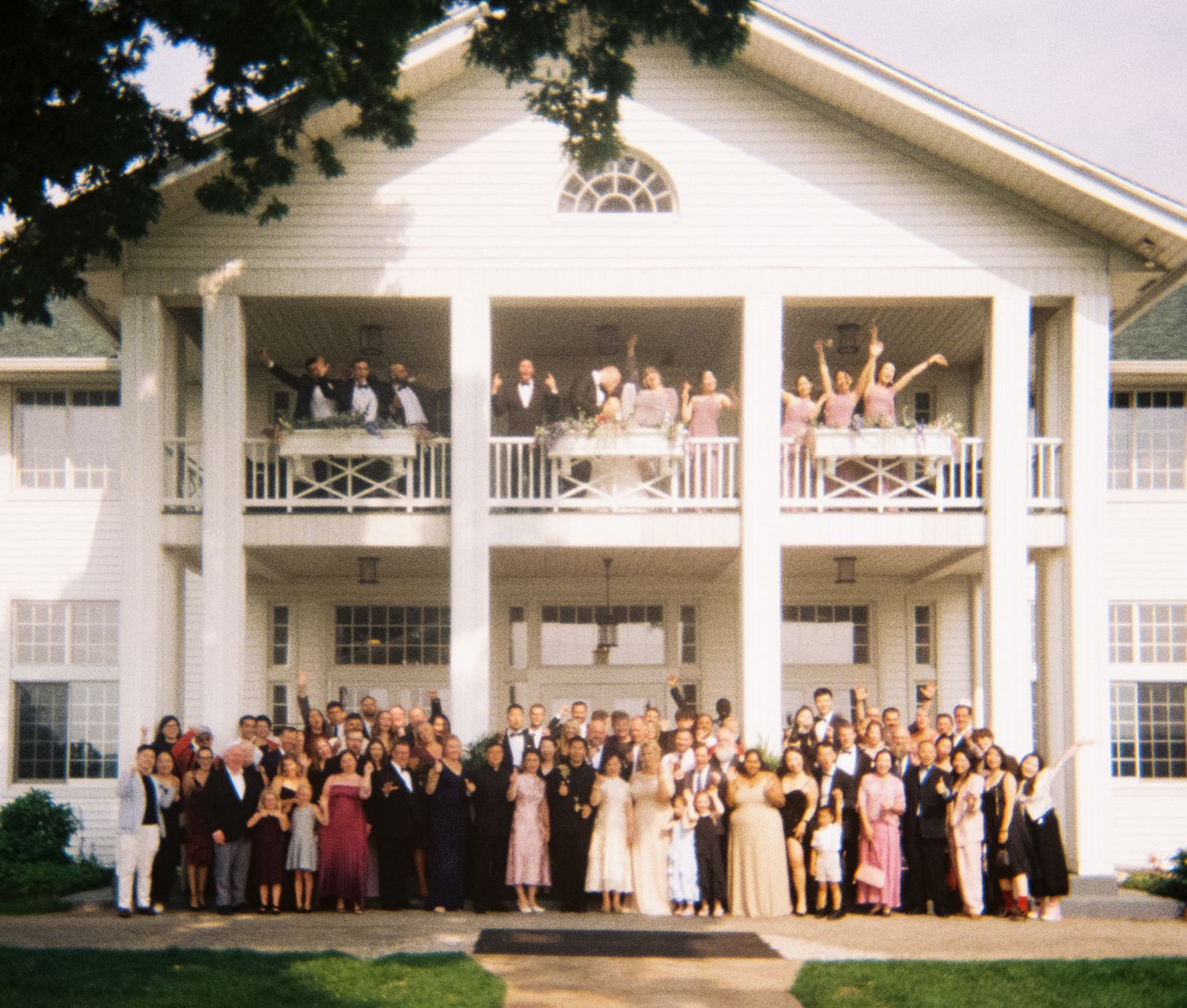 Large wedding party poses on white mansion porch for panoramic group photo in elegant formal attire.