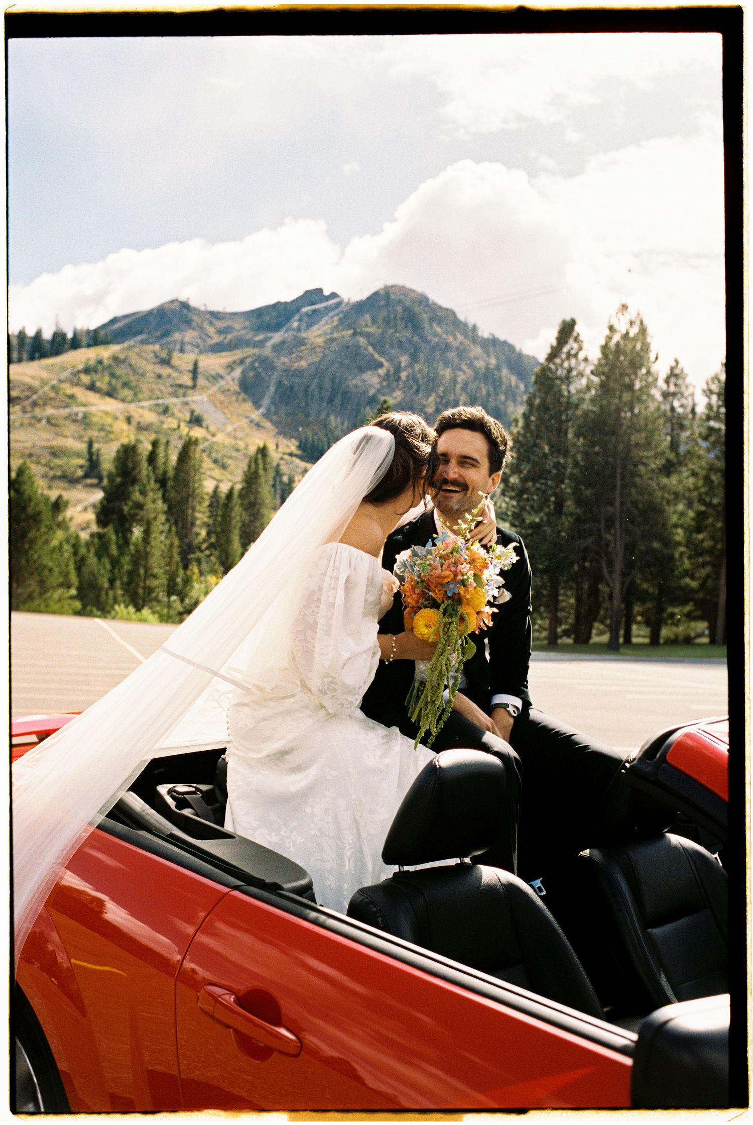 A wedding couple riding in a red convertible car with mountains and pine trees in the background.