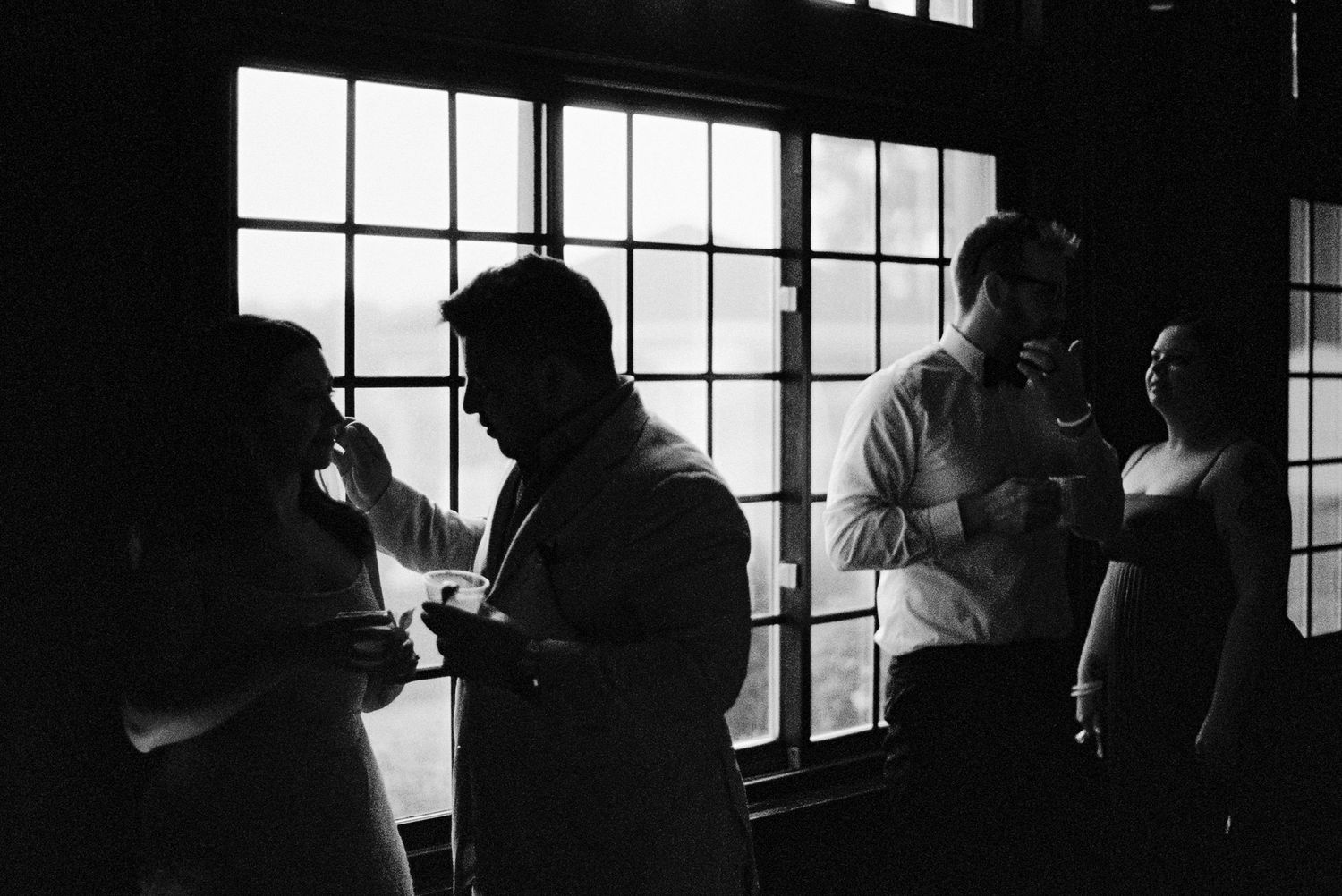 Silhouettes of wedding guests gathered by large windows in dramatic black and white photography.