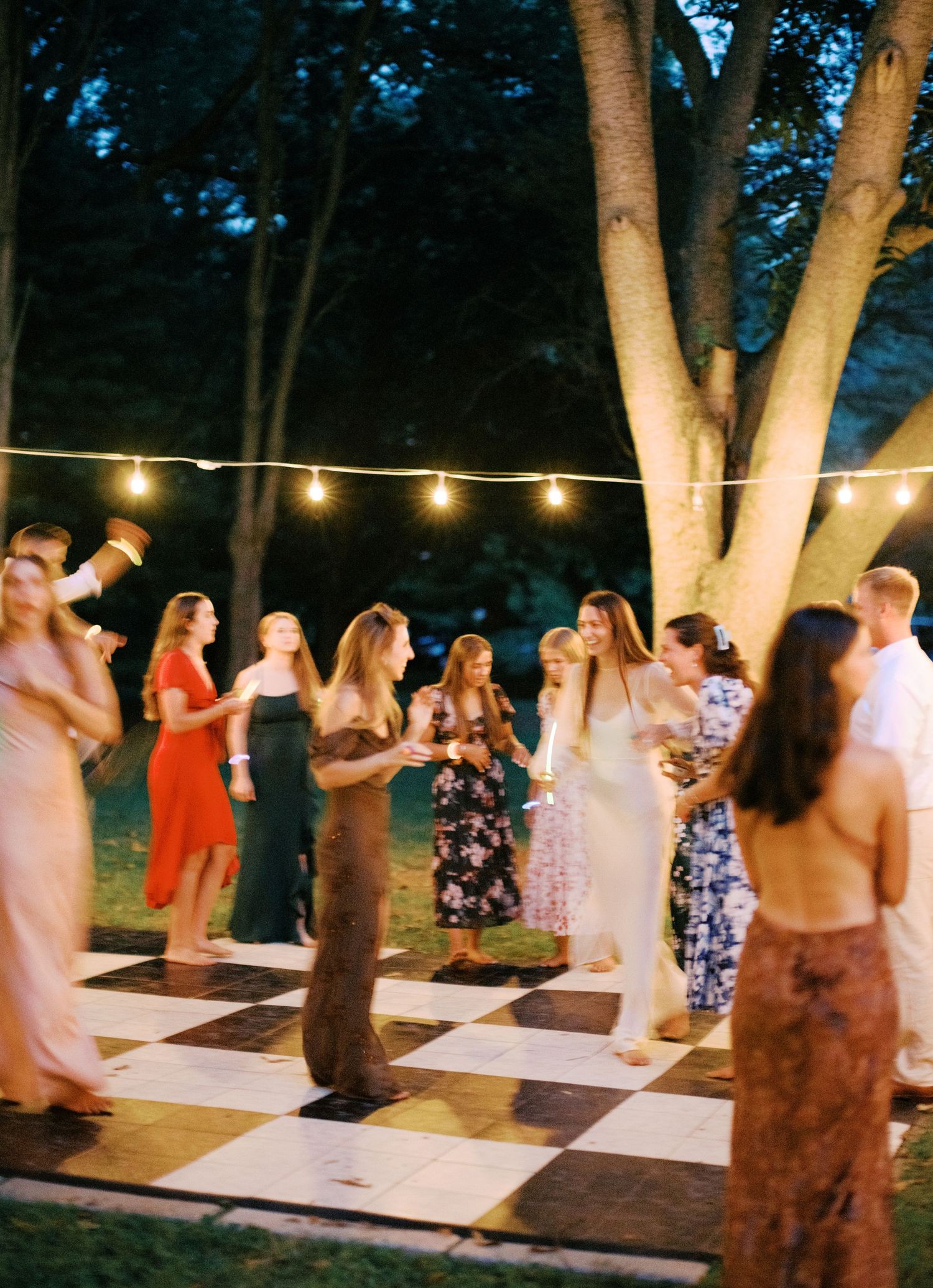 Outdoor wedding reception with string lights and guests dancing on a checkered dance floor.