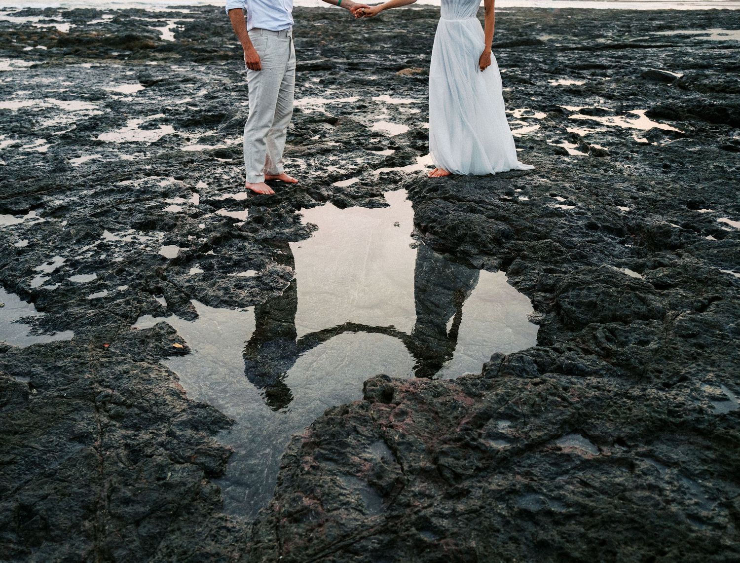 Wedding couple's reflection captured in dark volcanic rocks by the ocean.