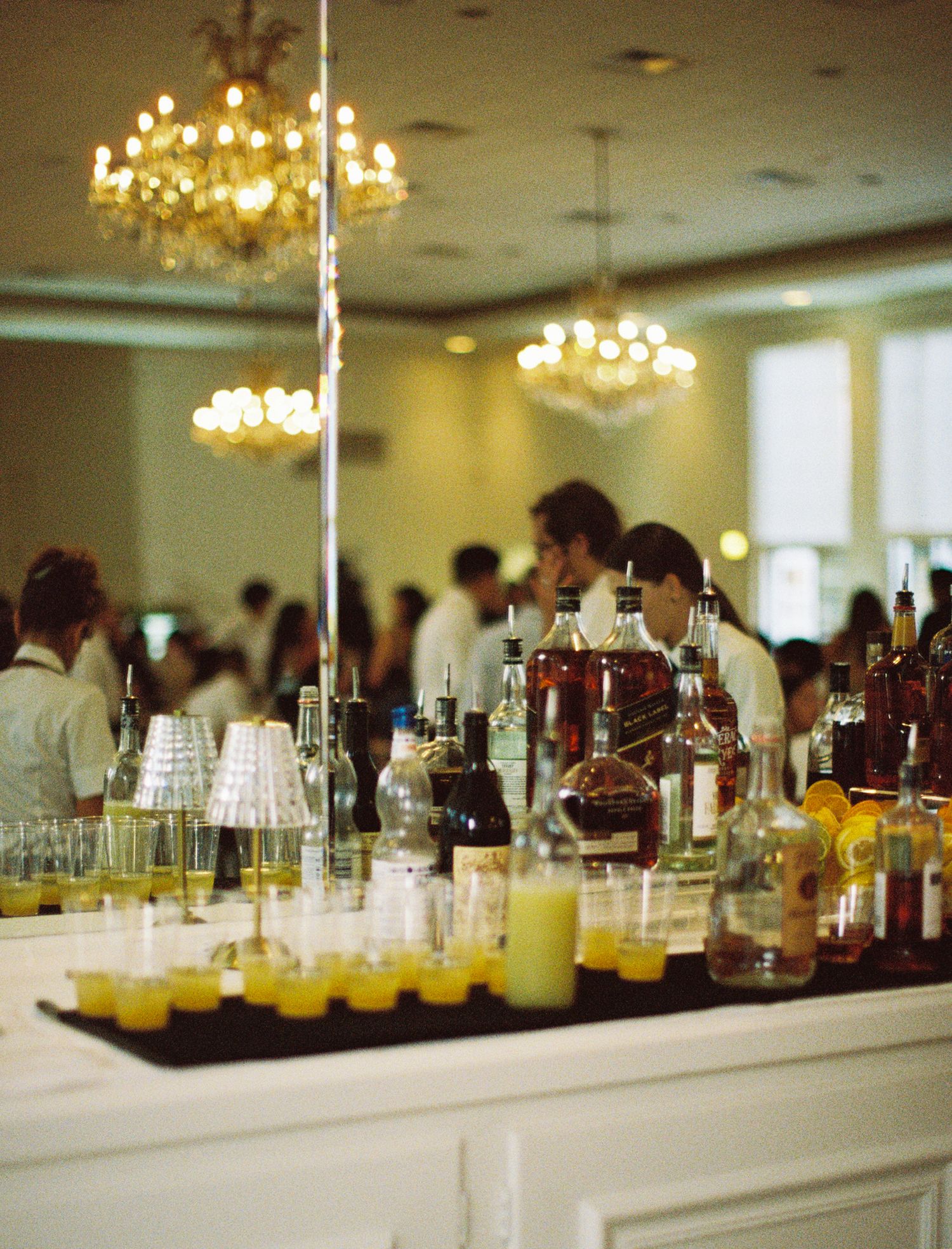 Long bar counter with various drinks and glassware under elegant chandeliers at an event venue.