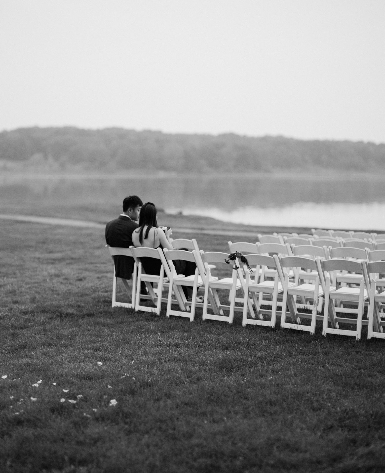Black and white photo of empty chairs by a lake after a wedding ceremony.