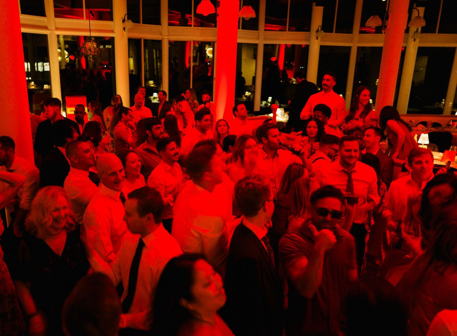 A crowded nightclub bathed in red lighting shows people dancing and socializing in a dark atmospheric space.