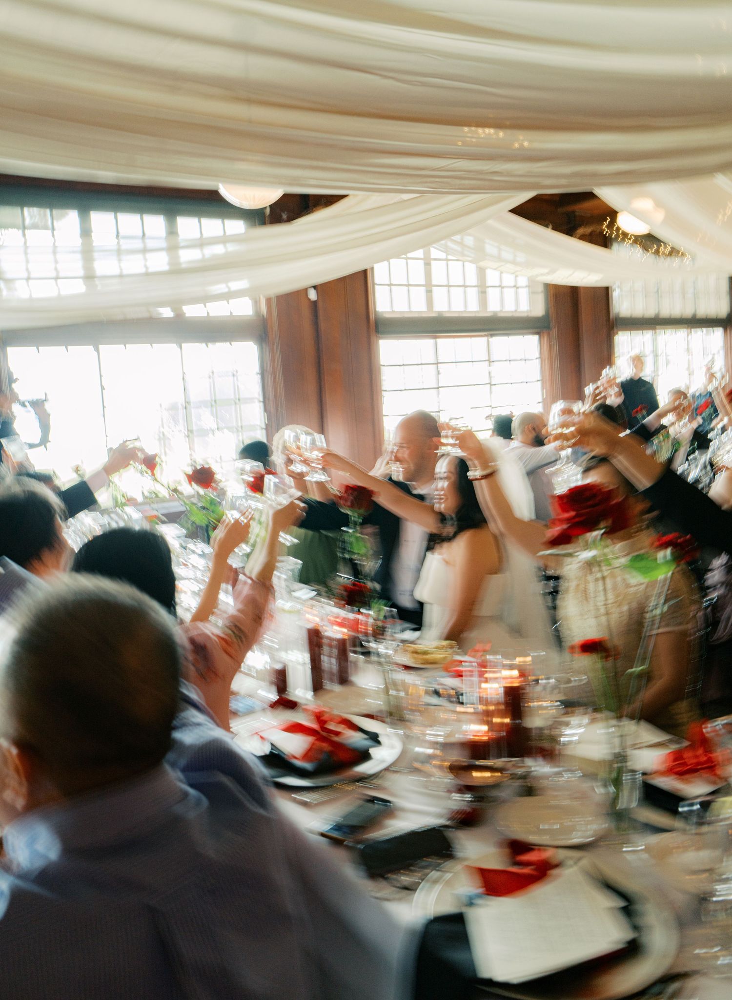 Motion-blurred image of wedding guests raising glasses in celebration under white draped ceiling.
