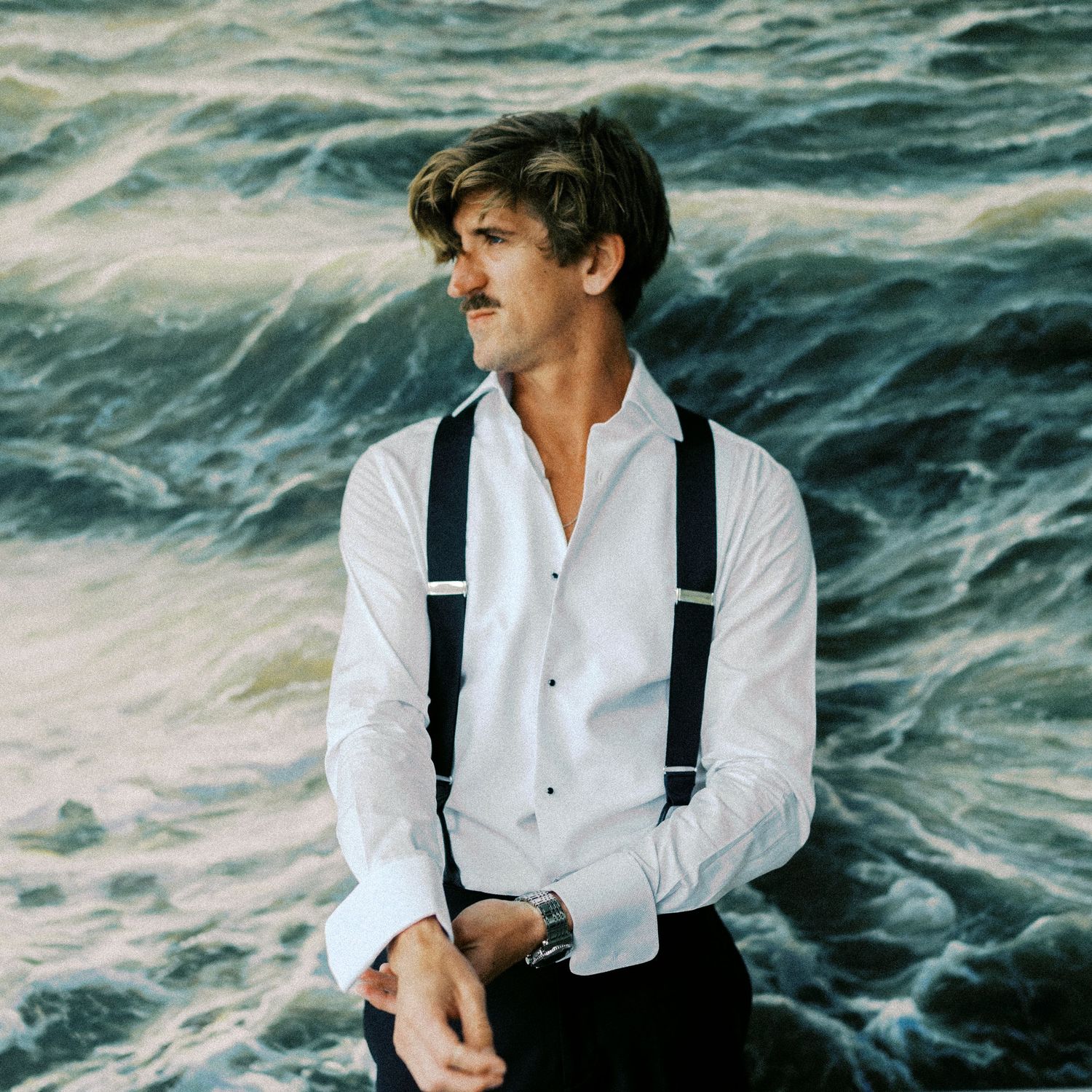 Person in white shirt and suspenders gazes across ocean waves in dramatic black and white photograph.
