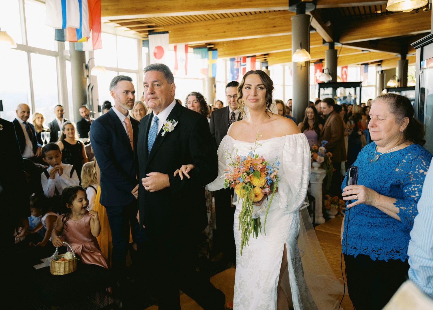 A wedding ceremony takes place in a bright indoor venue with guests gathered as the bride holds a vibrant sunflower bouquet.