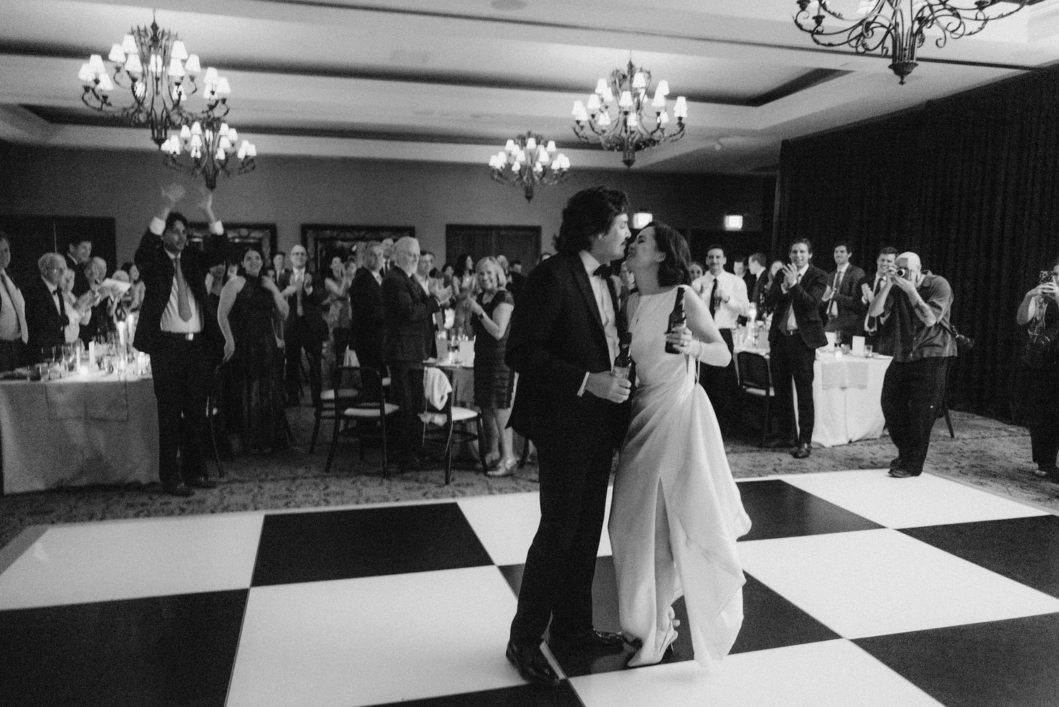 A couple shares their first dance on a black and white checkered floor at an elegant reception with crystal chandeliers.