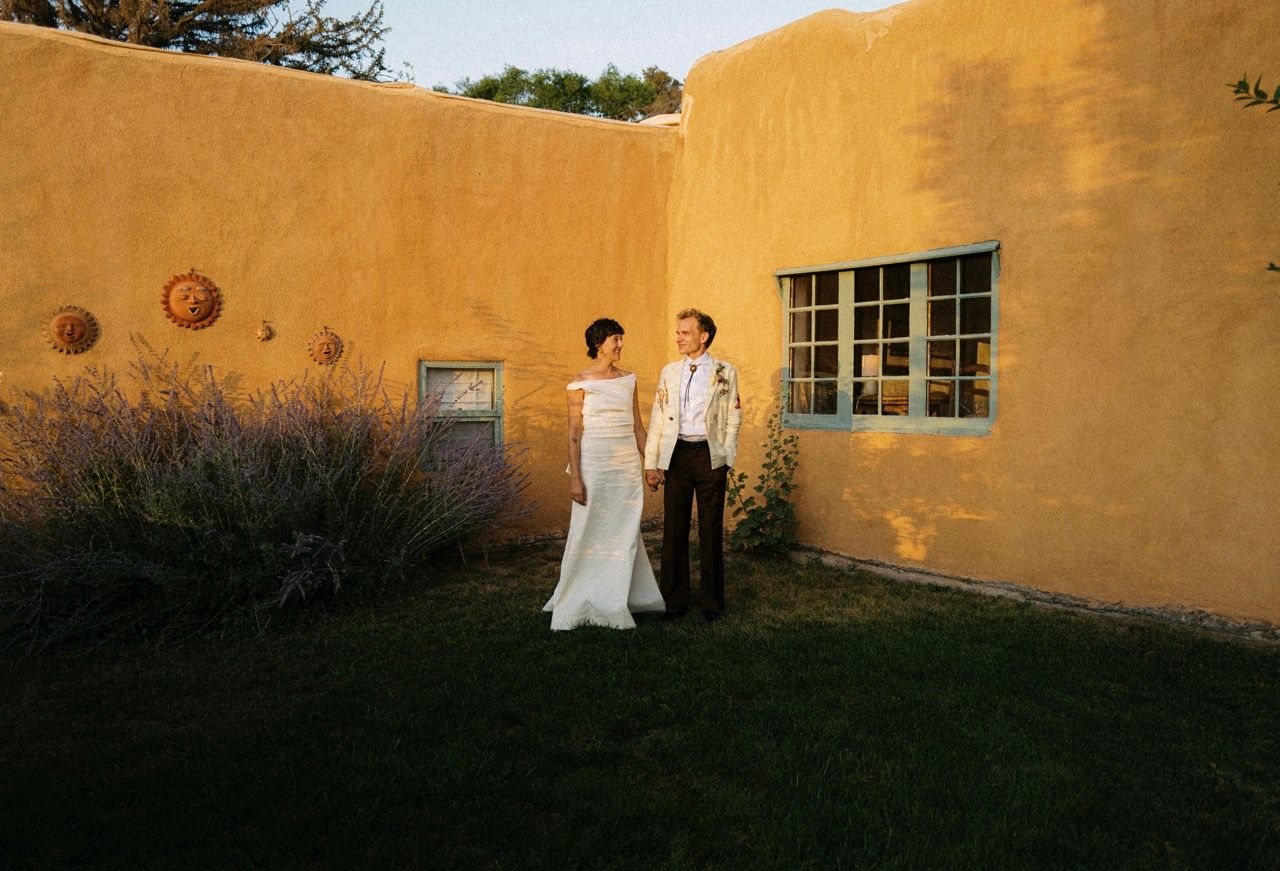 A person in white stands against a golden yellow adobe wall with windows during sunset.
