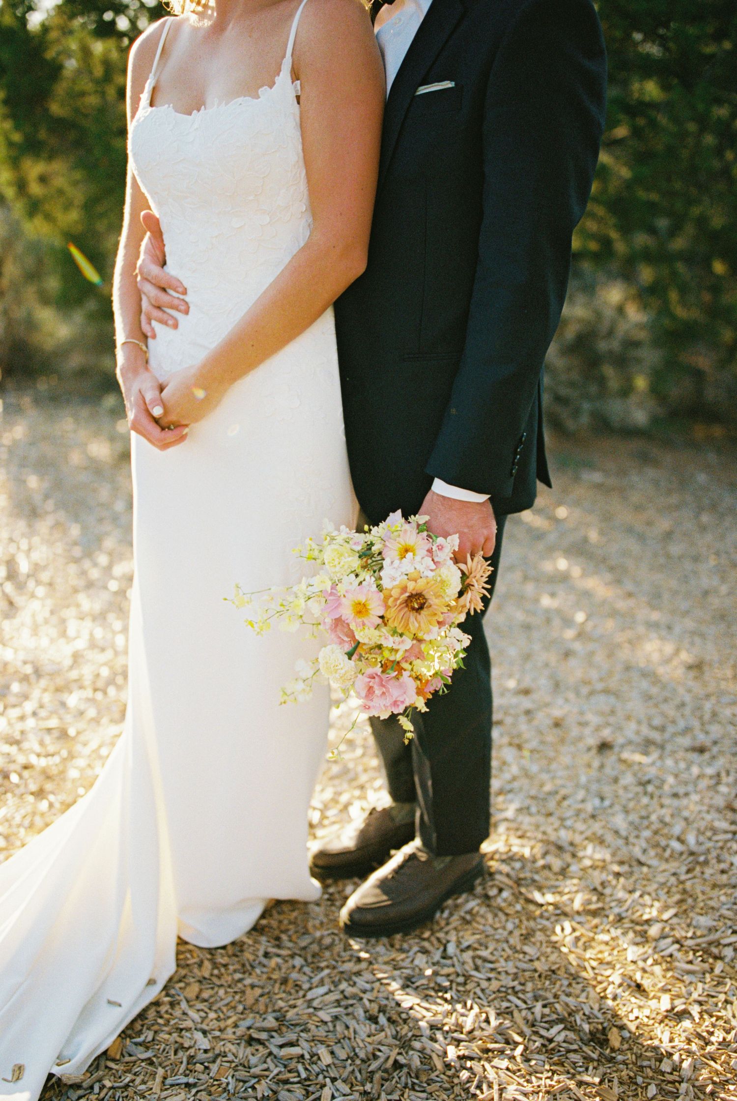 Close-up of bride and groom's feet standing together on gravel path with sunlight filtering through trees.