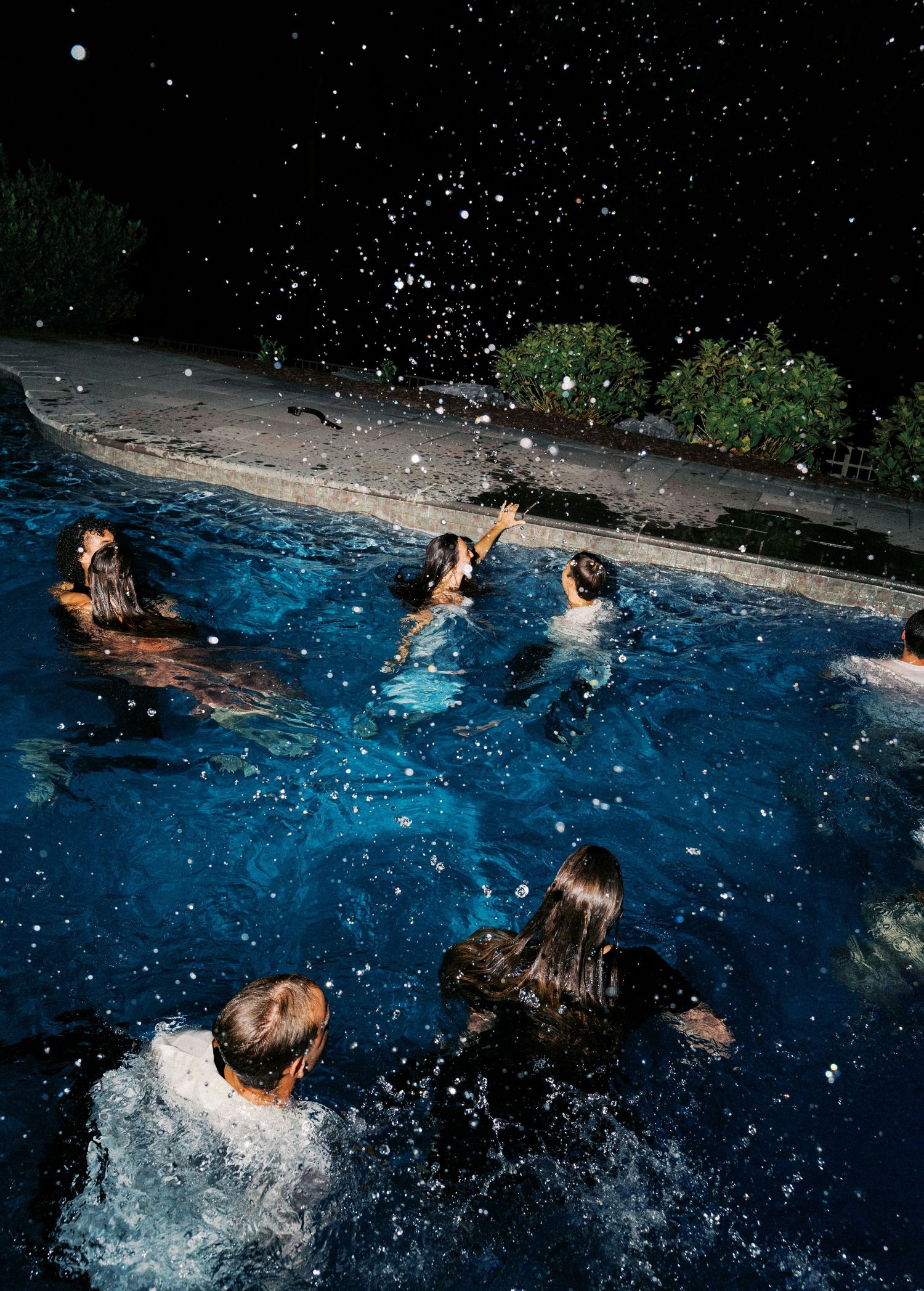 People splashing and playing in a blue swimming pool at night with water droplets sparkling.