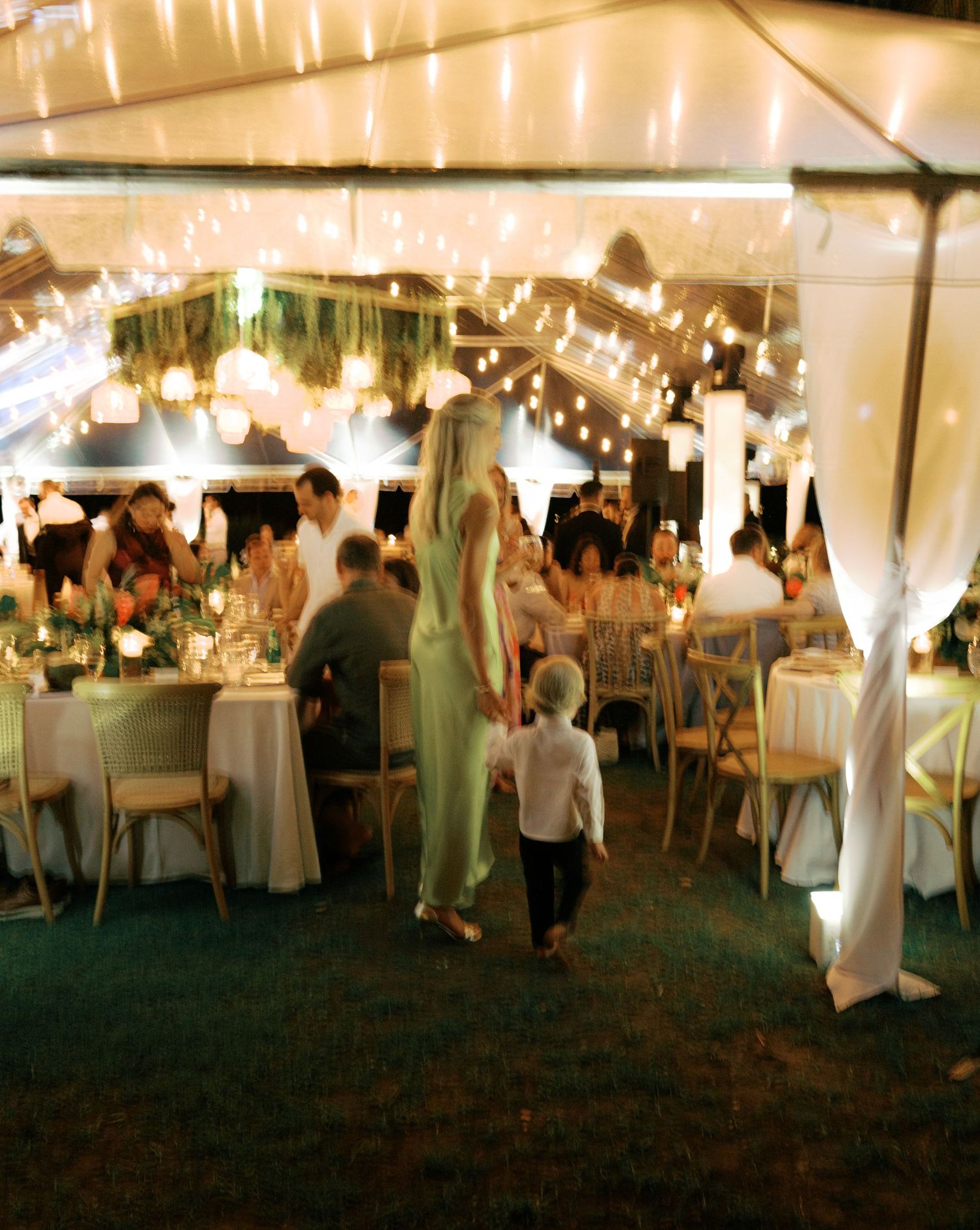 People mingle and socialize under a white tent at an evening reception with string lights creating a warm ambiance.