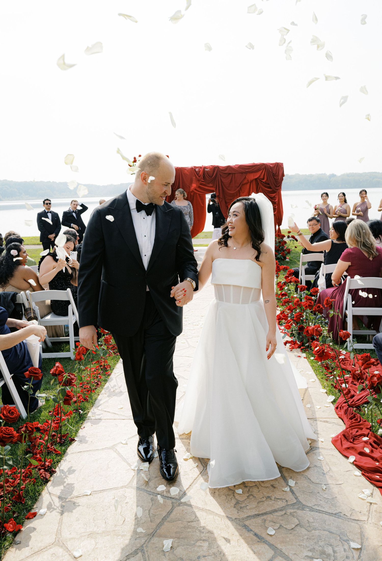 Wedding couple walks down stone path lined with red flowers during outdoor ceremony.