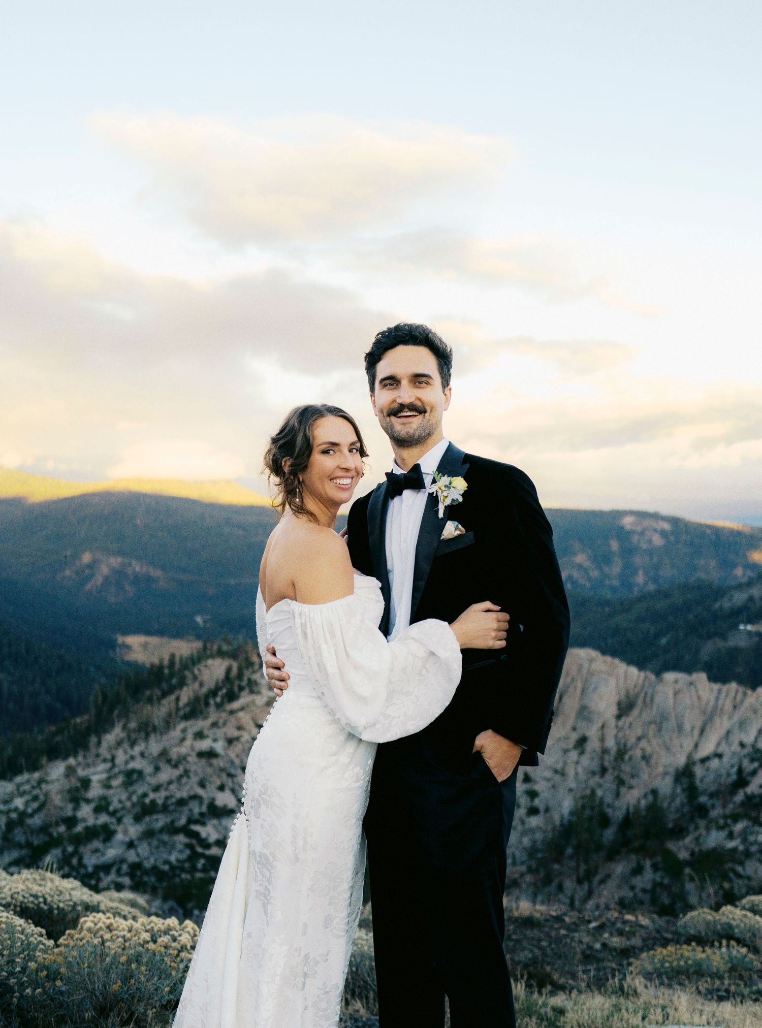 A couple in wedding attire embraces on a mountaintop with dramatic cliffs and sunset sky in the background.