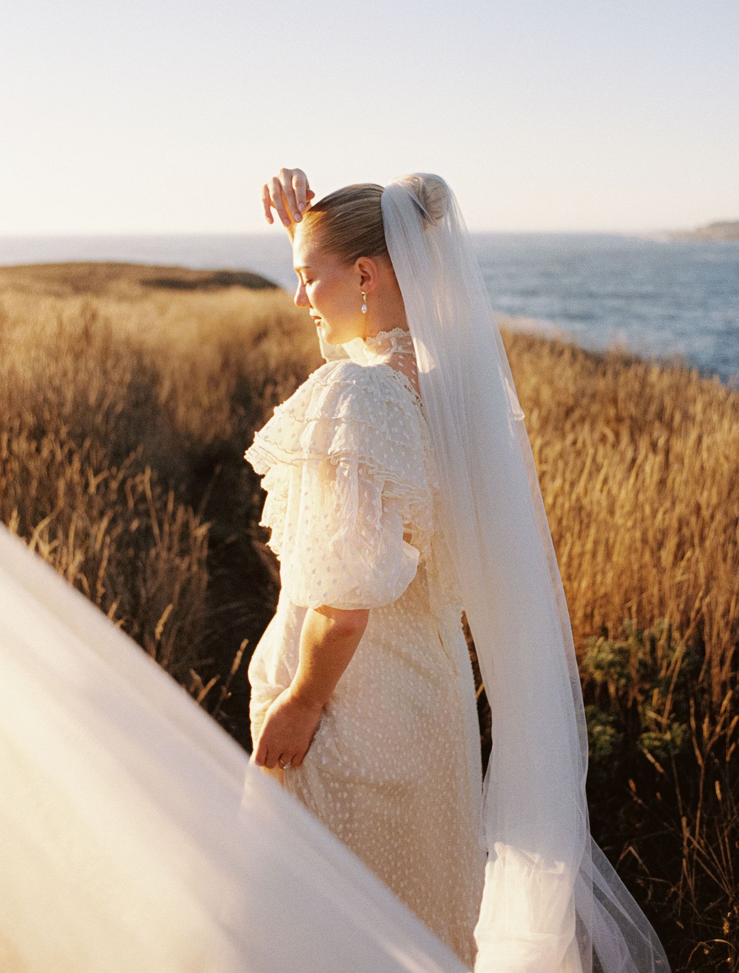 A white wedding dress with veil flows in the golden sunlight against a backdrop of tall dried grass and ocean views.