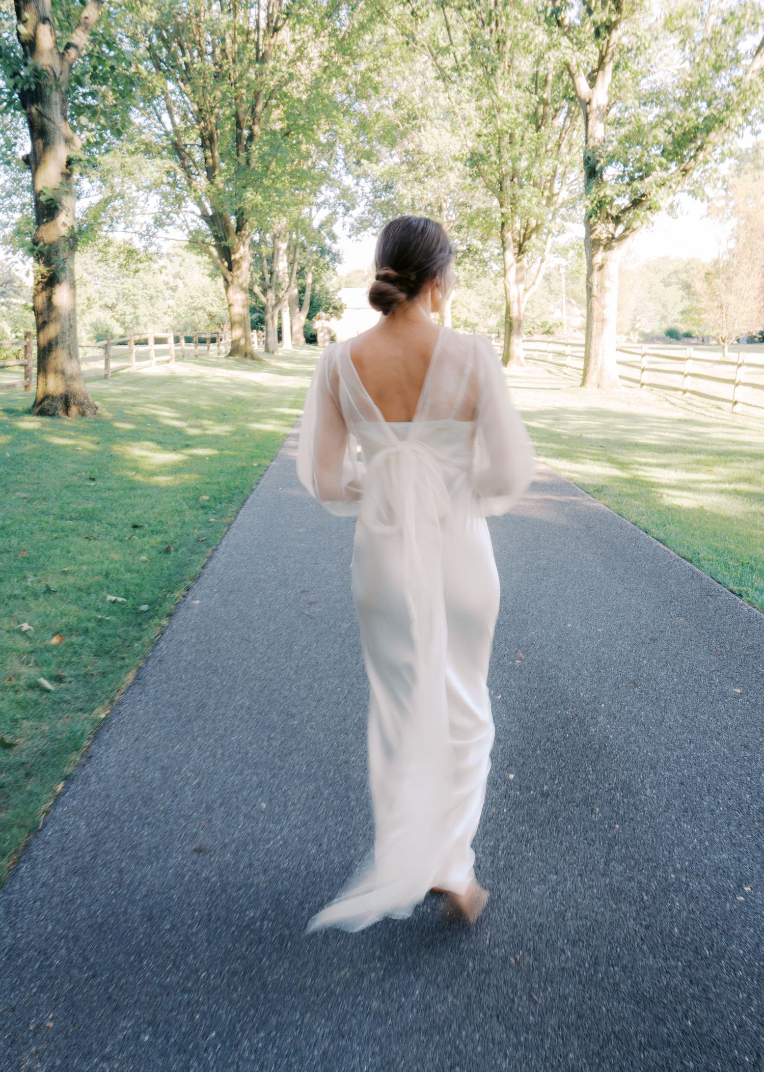 A flowing white dress twirls on a paved path surrounded by green trees in an outdoor setting.