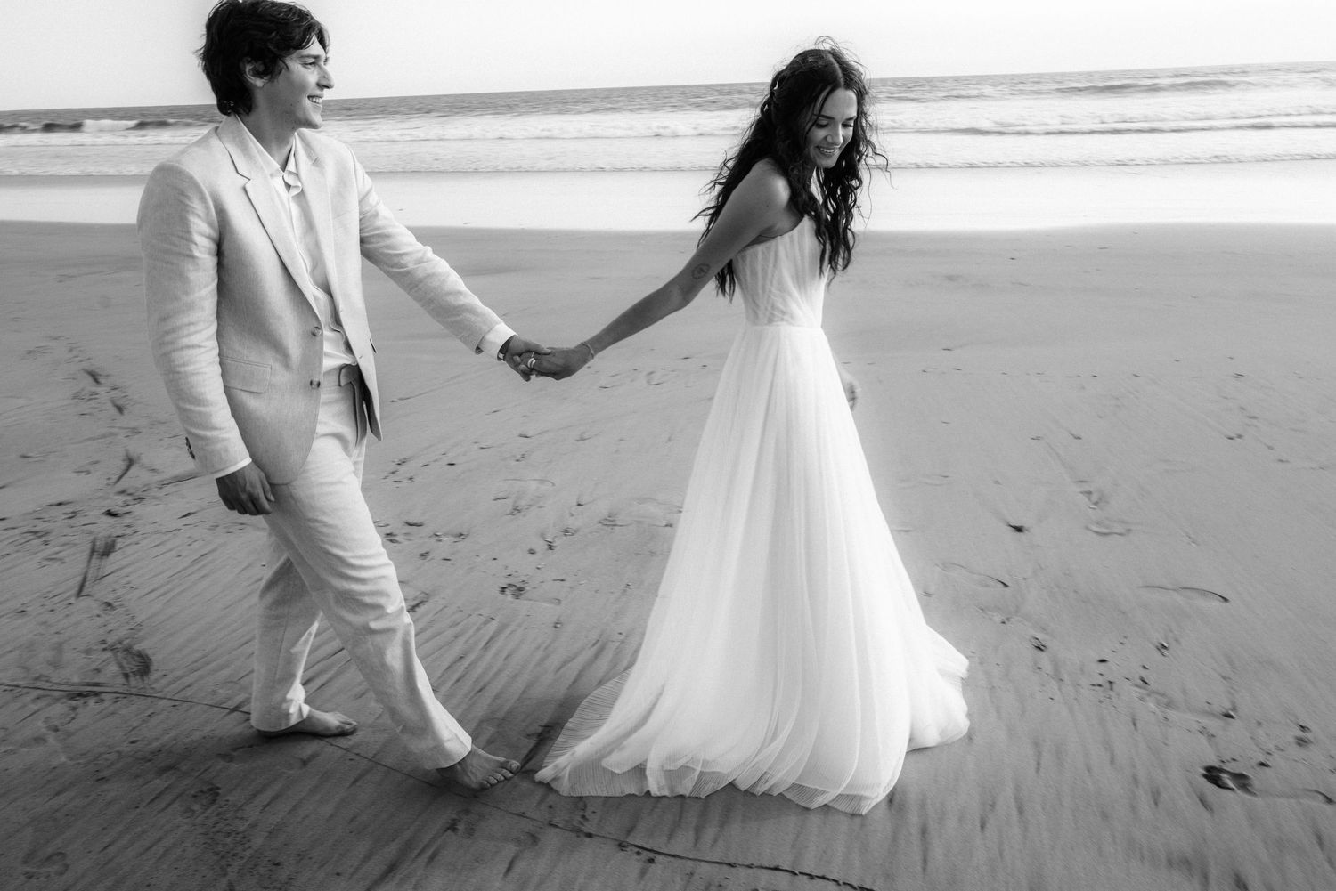 Newlywed couple walking hand in hand along a sandy beach in black and white.