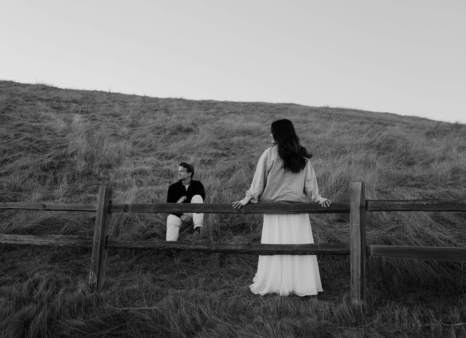 Two people sit on a wooden fence overlooking a grassy hillside in black and white photography.