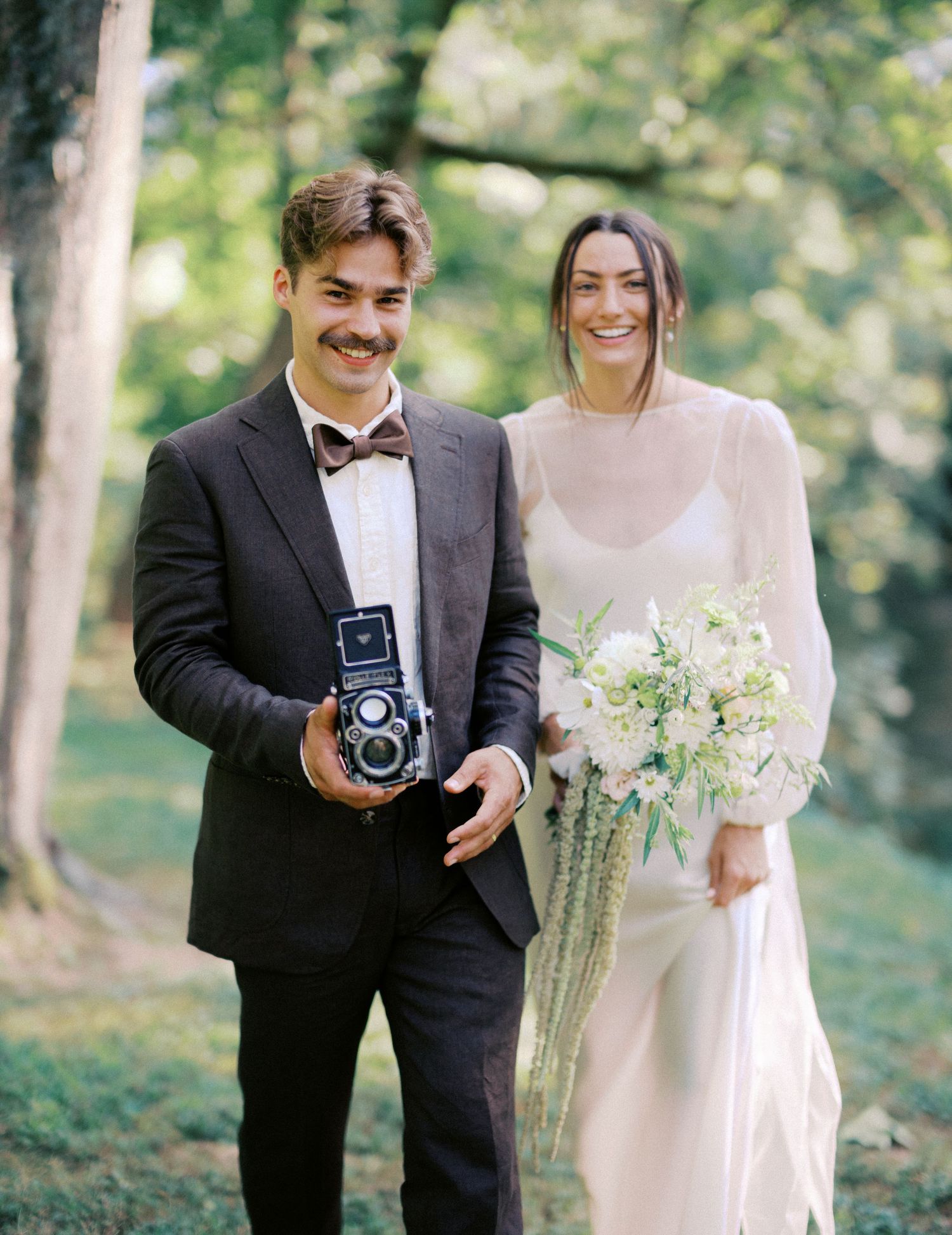 A couple poses with vintage cameras during their wedding photoshoot in a wooded setting.