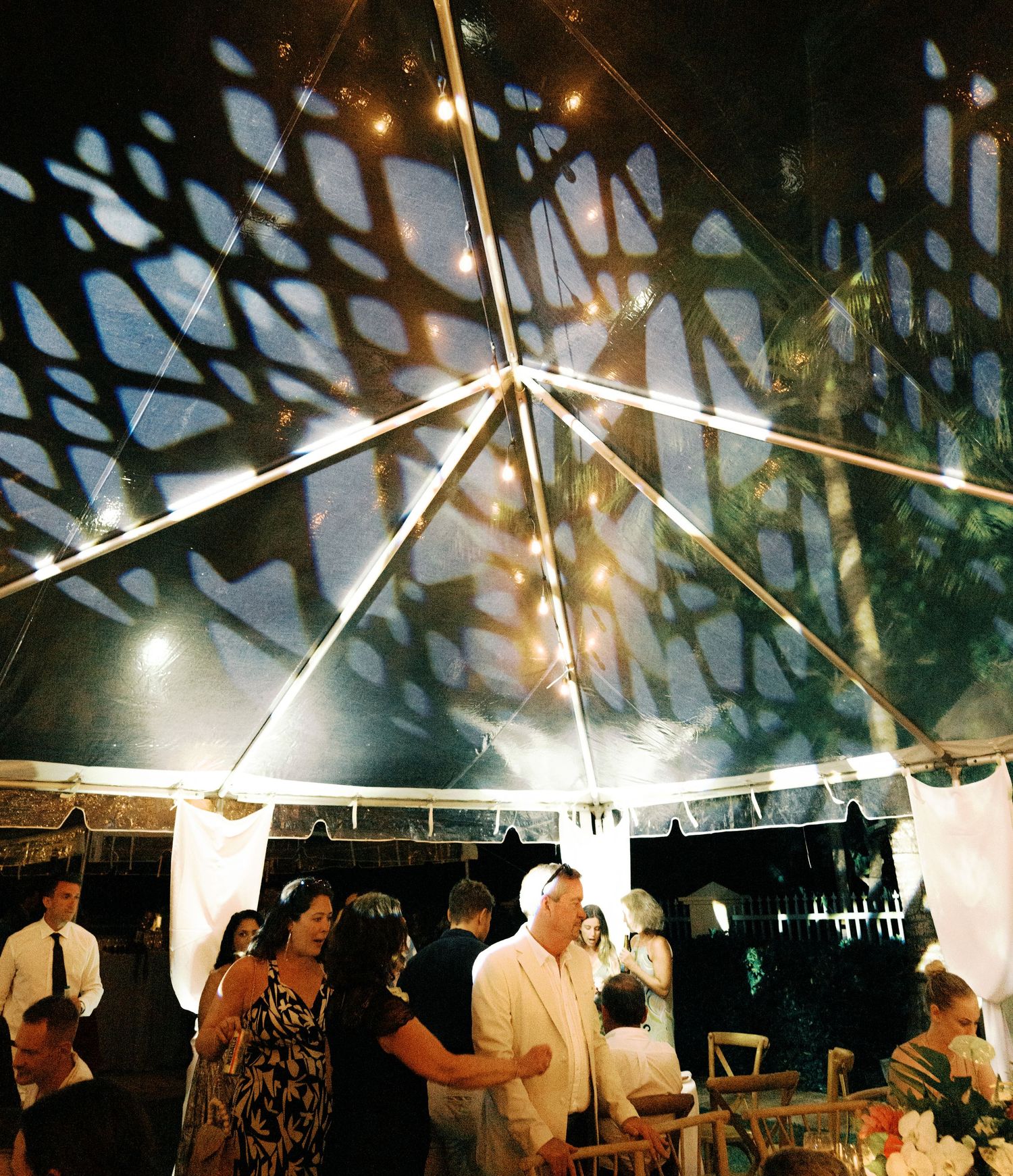 Wedding guests dance and celebrate under a white tent illuminated by string lights during an evening reception.