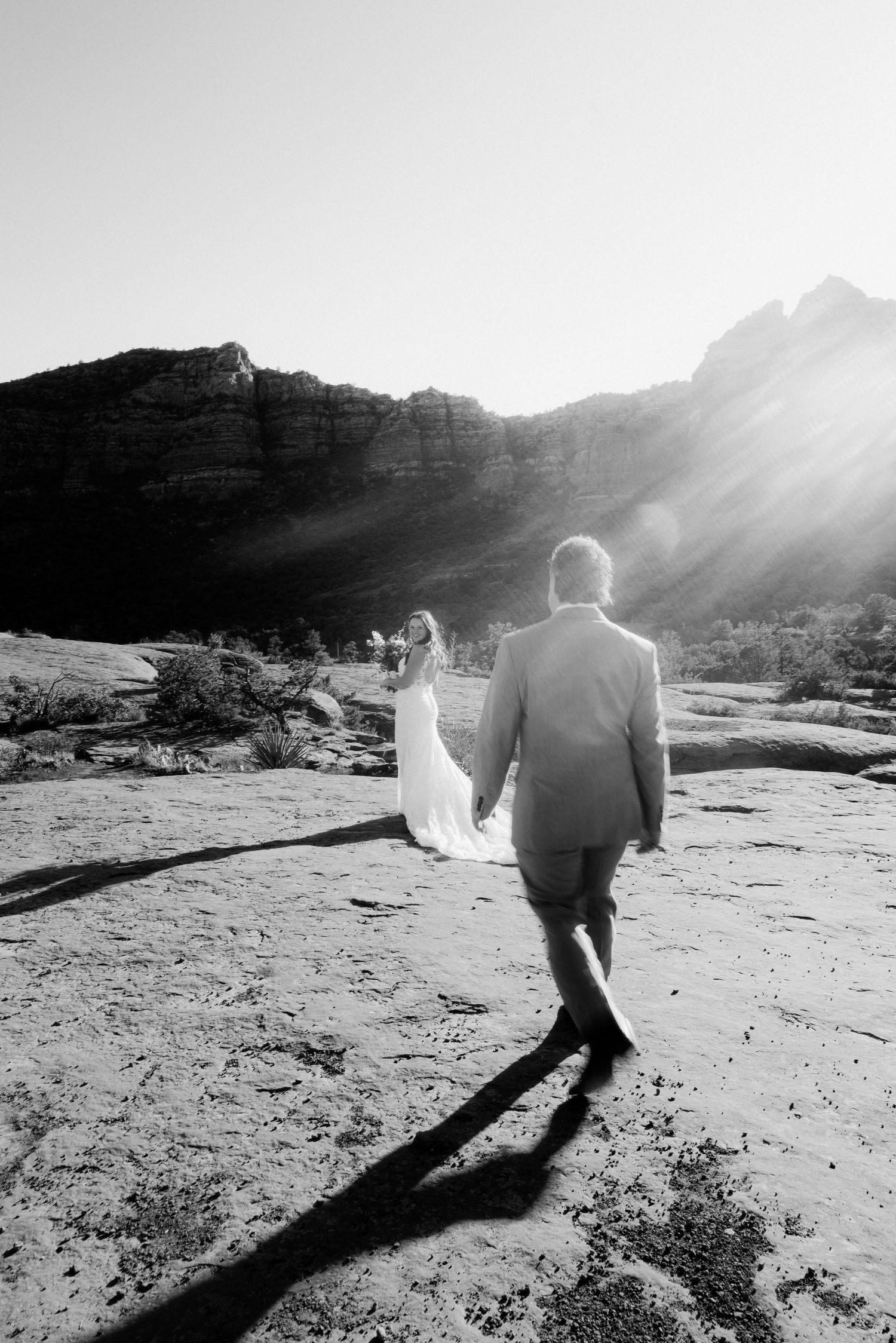 A couple walks together casting long shadows across desert rocks as sunlight streams through mountain peaks in black and white.