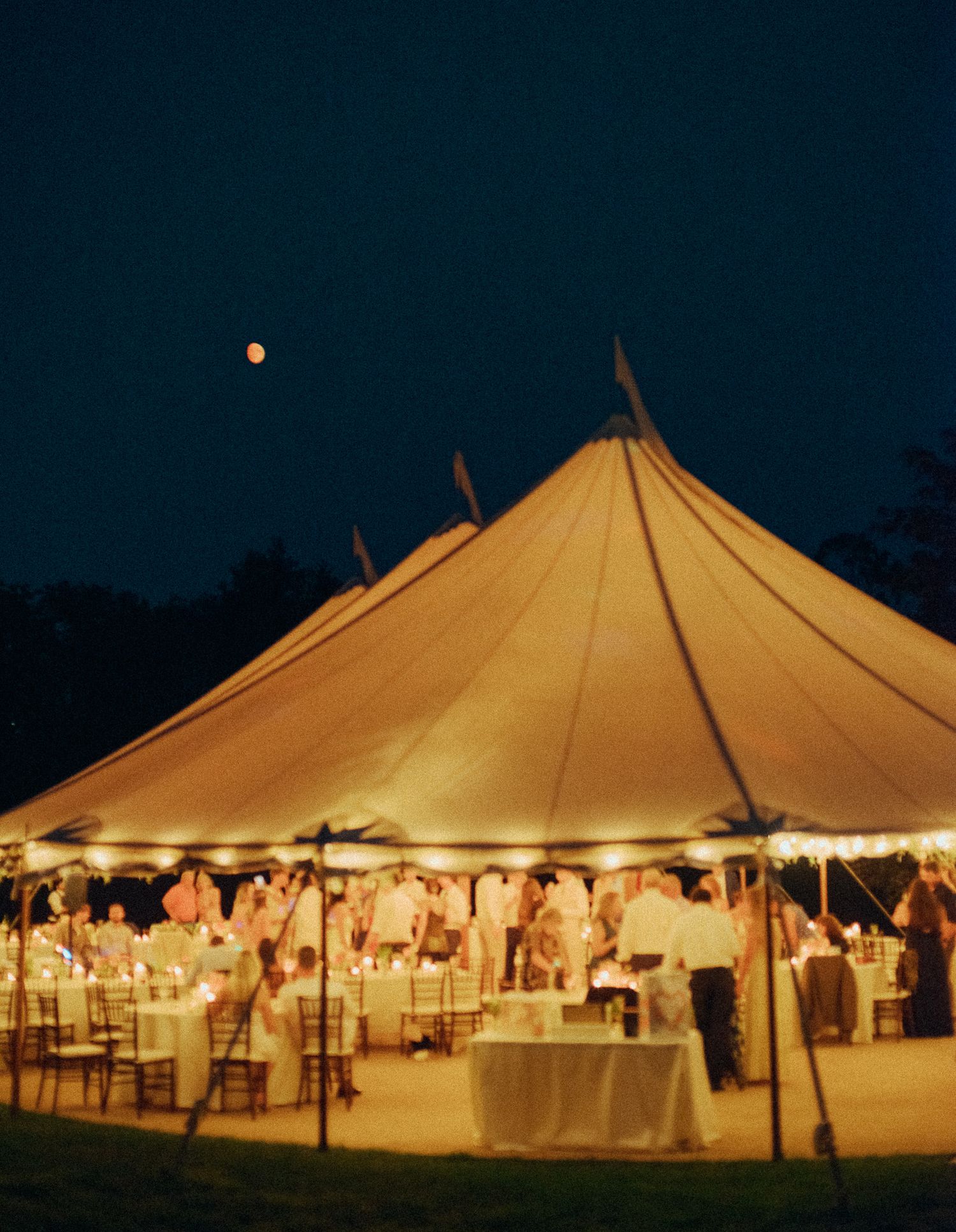 Illuminated white wedding tents glowing at dusk with string lights creating an elegant outdoor reception atmosphere.