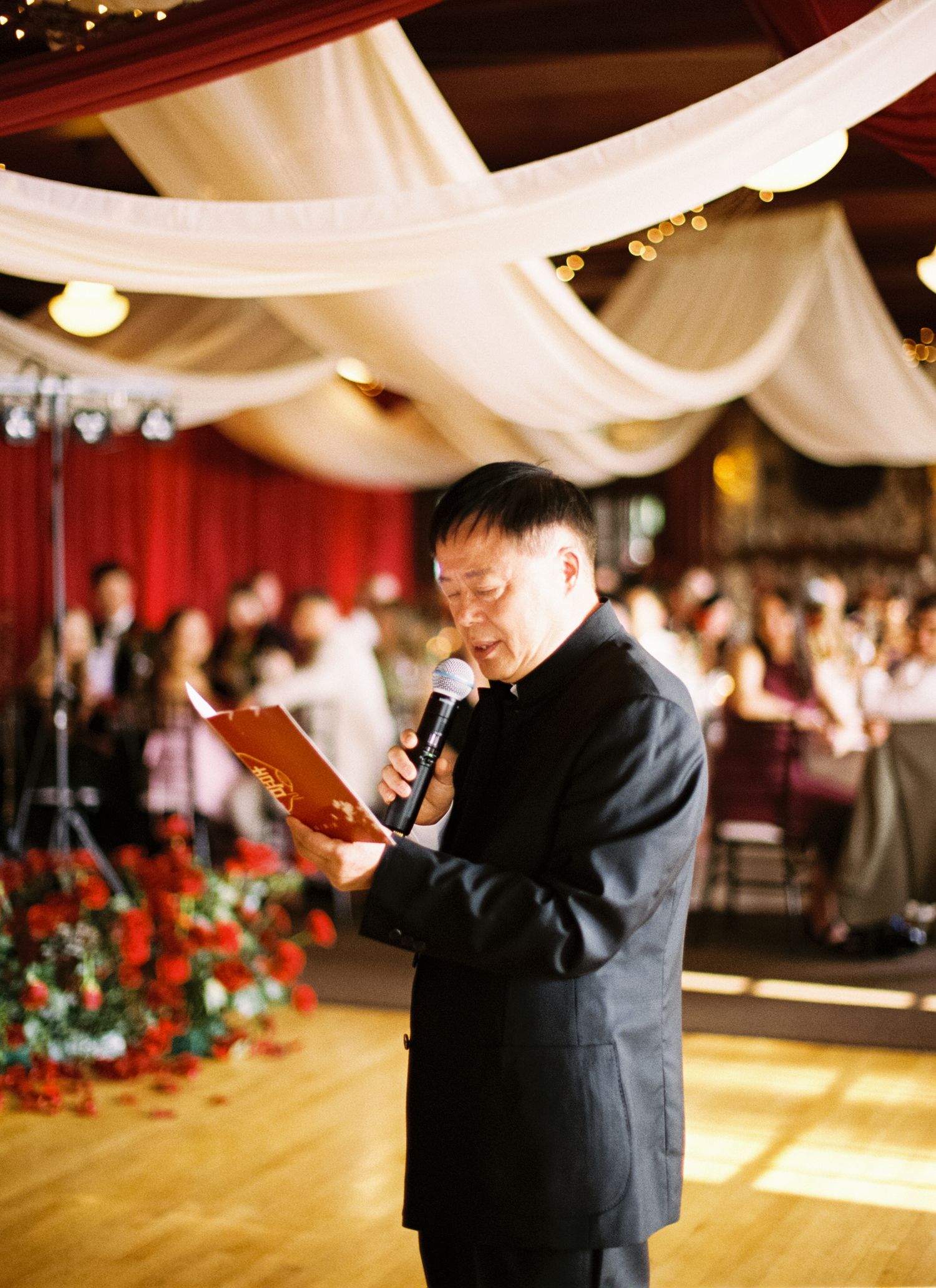 A speaker in formal attire reads from a document under elegant white ceiling drapes at a wedding reception.