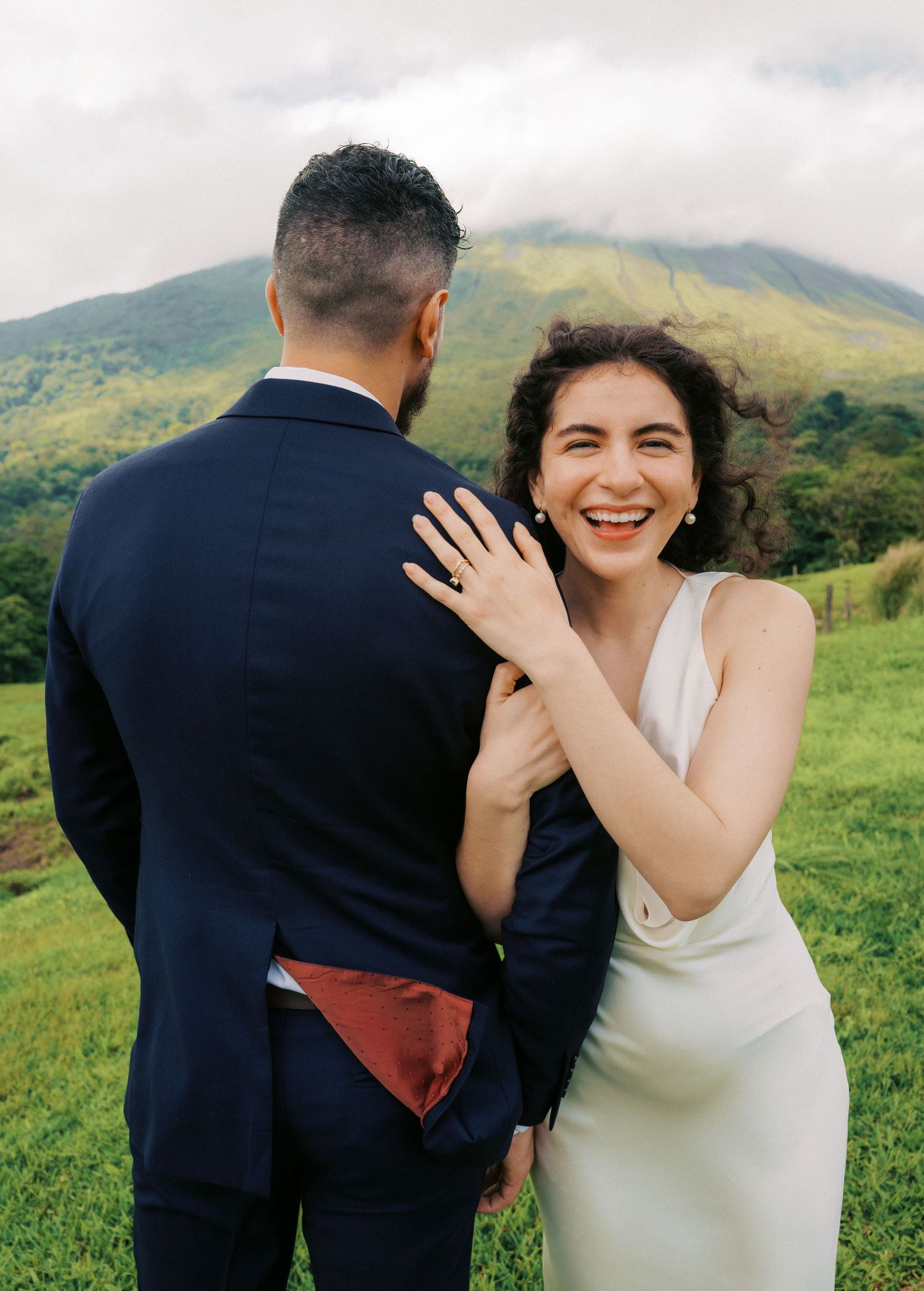 A couple in wedding attire share a joyful moment together on a green hillside with mountains in the background.