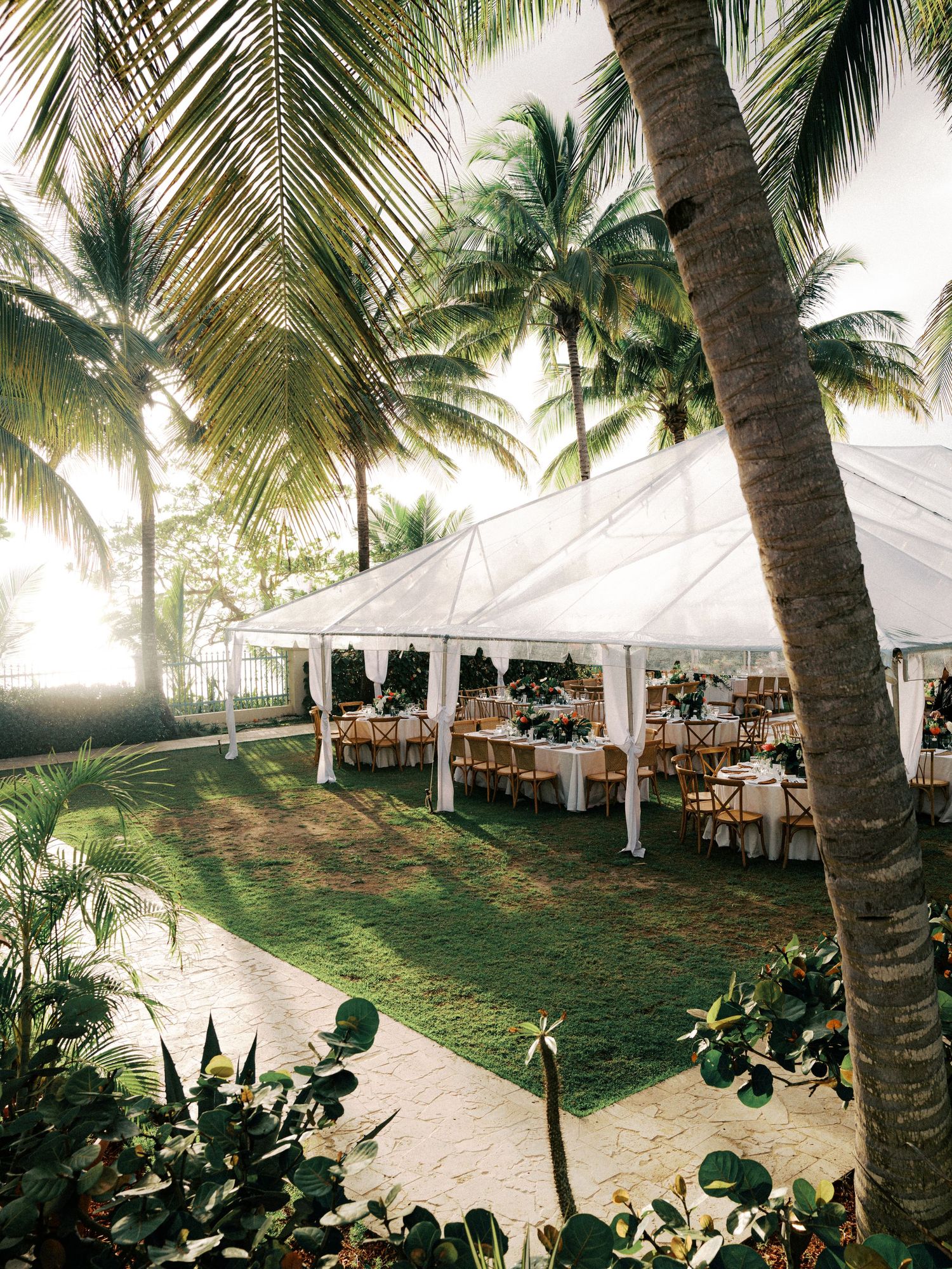 Elegant white wedding reception tent setup on a tropical beach with palm trees and ocean views at sunset.