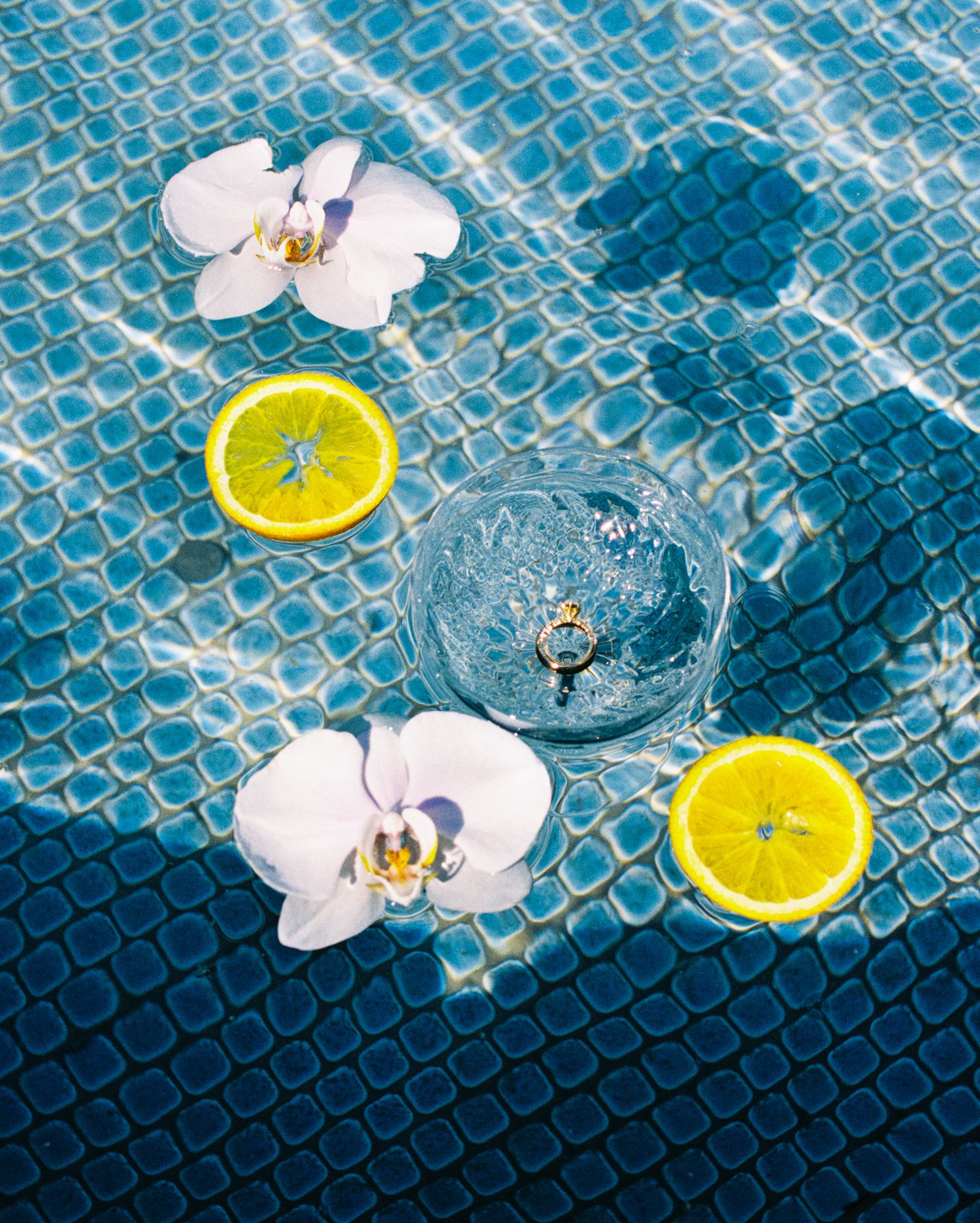 White orchid flowers and lemon slices float on rippling blue swimming pool water with mosaic tile pattern beneath.