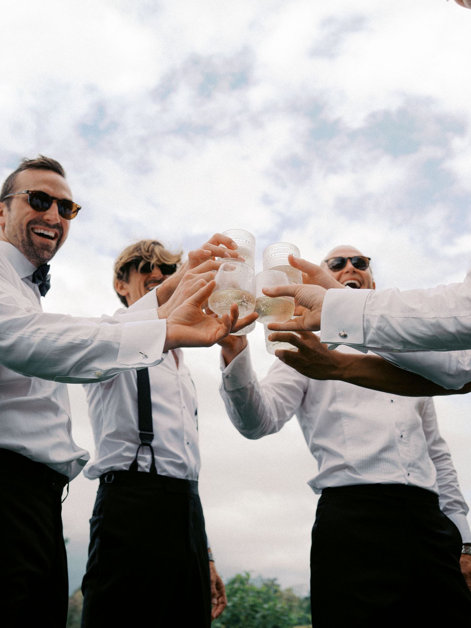 Groomsmen in white shirts and black pants perform a synchronized celebratory toast outdoors under cloudy skies.