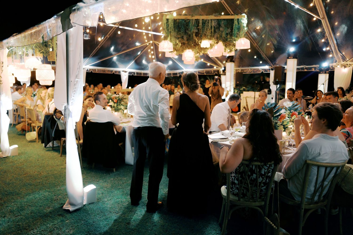 Guests dance and celebrate at an outdoor evening wedding reception under string lights and a white tent.