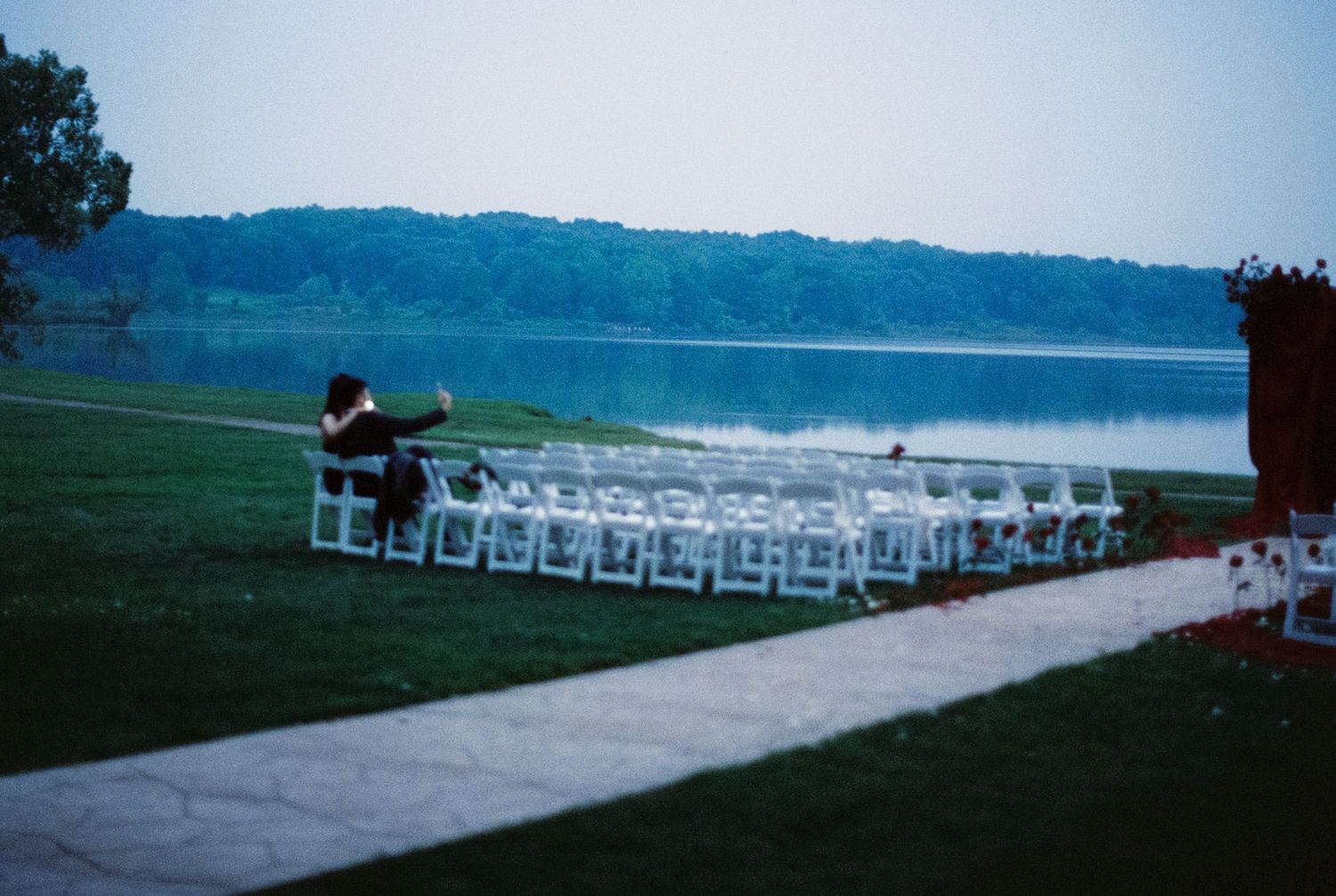 White chairs arranged for outdoor wedding ceremony on grassy lawn overlooking serene lake at dusk.