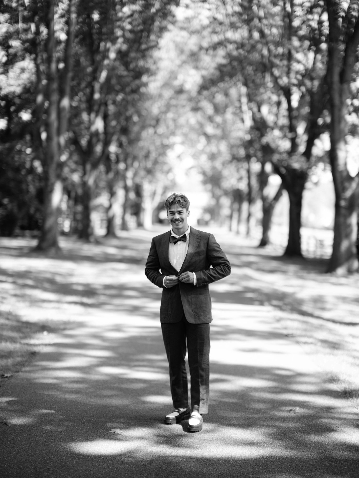 A black and white photo series shows a person in formal attire walking and dancing playfully in a tree-lined path.