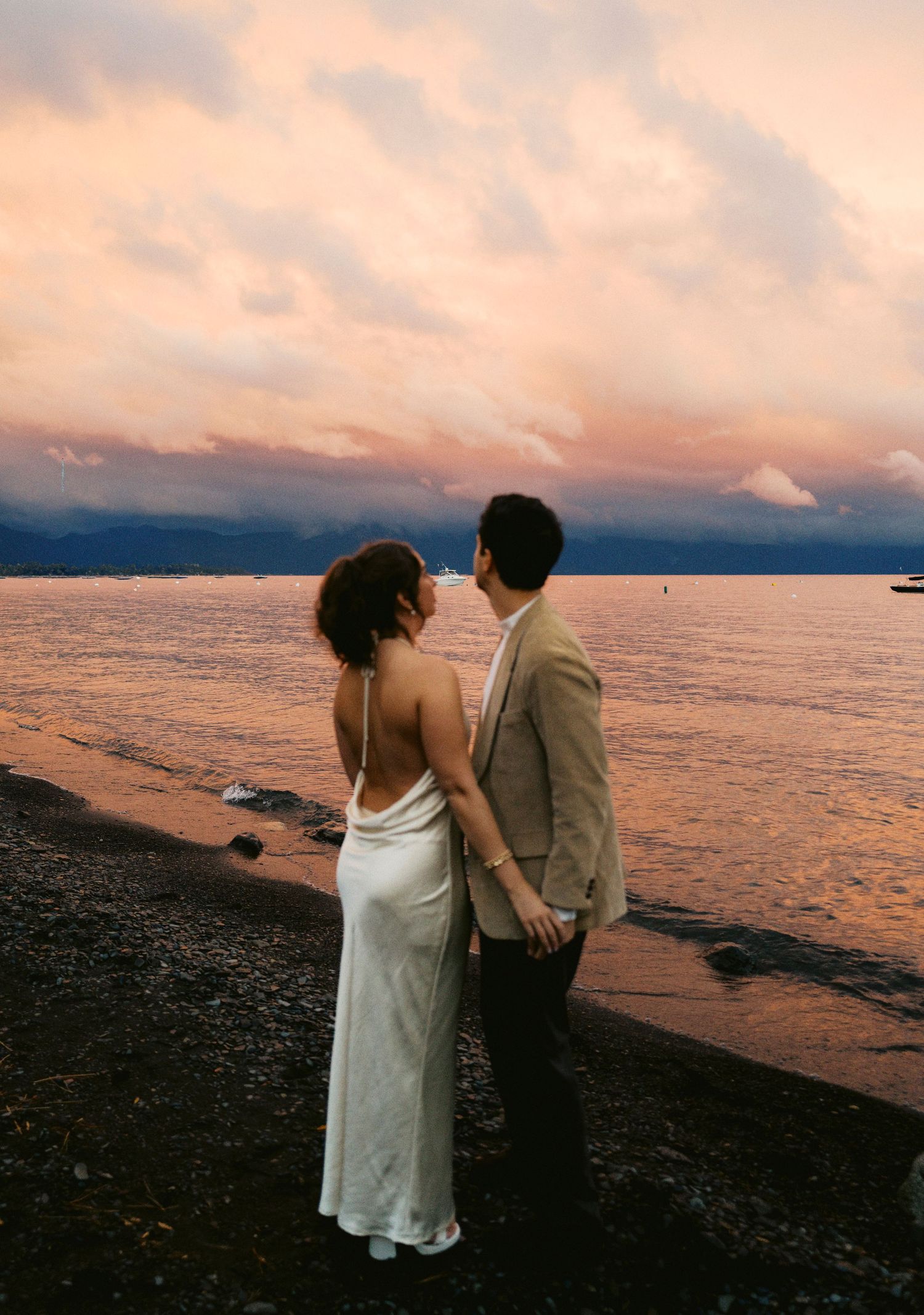 A couple shares a romantic moment on a beach at sunset with dramatic pink and orange clouds.