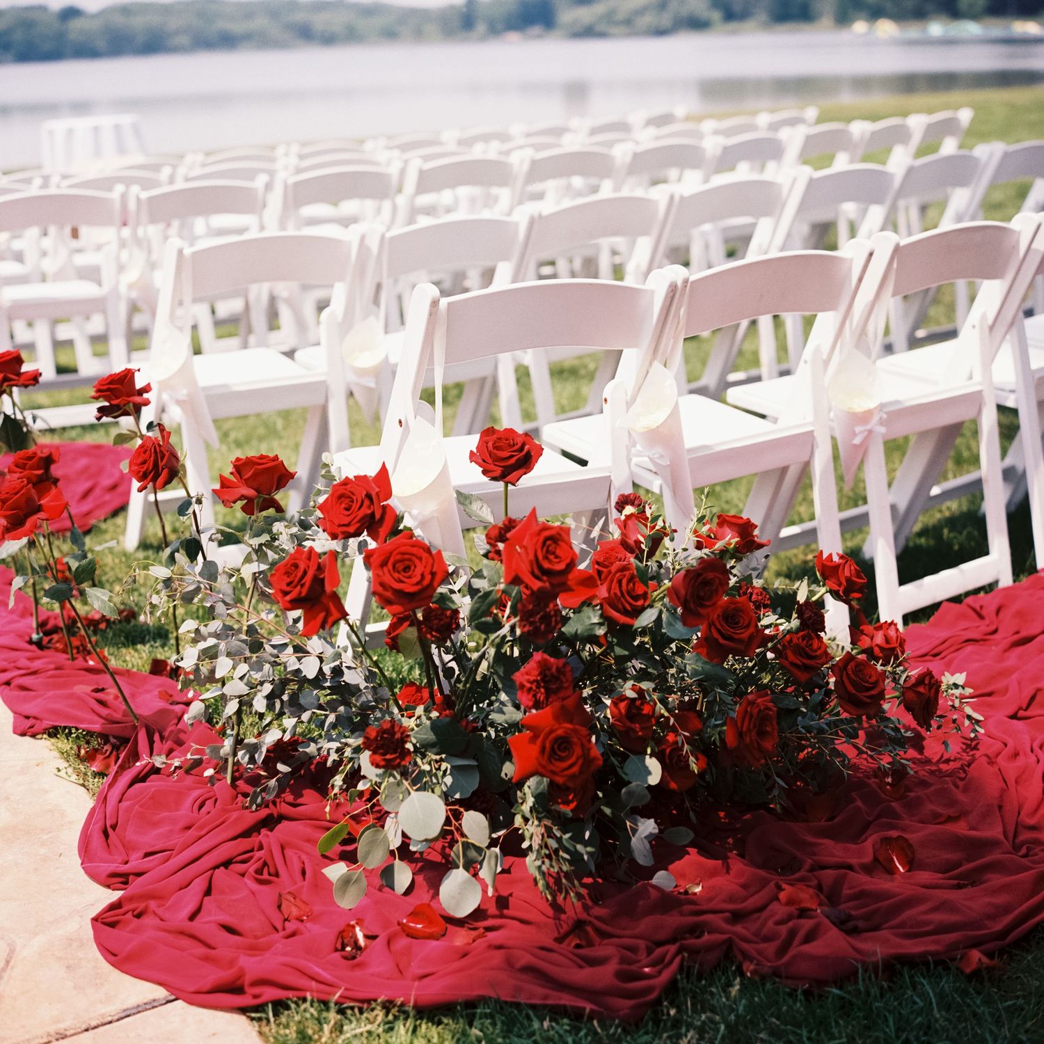 White folding chairs set up for outdoor wedding ceremony with red roses and fabric decorating the aisle.