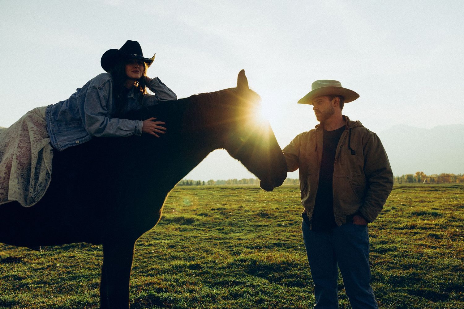 Two people in cowboy hats stand in a grassy field at sunset with dramatic silhouettes and lens flare.
