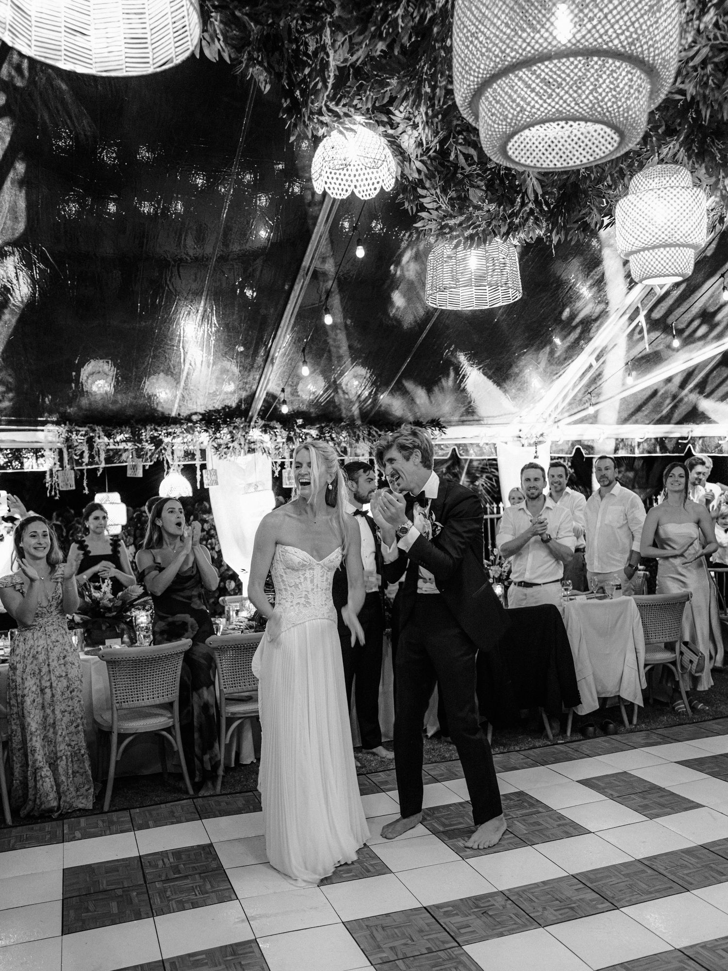 Black and white photo of a wedding reception with decorative lanterns overhead and guests dancing on a checkered floor.