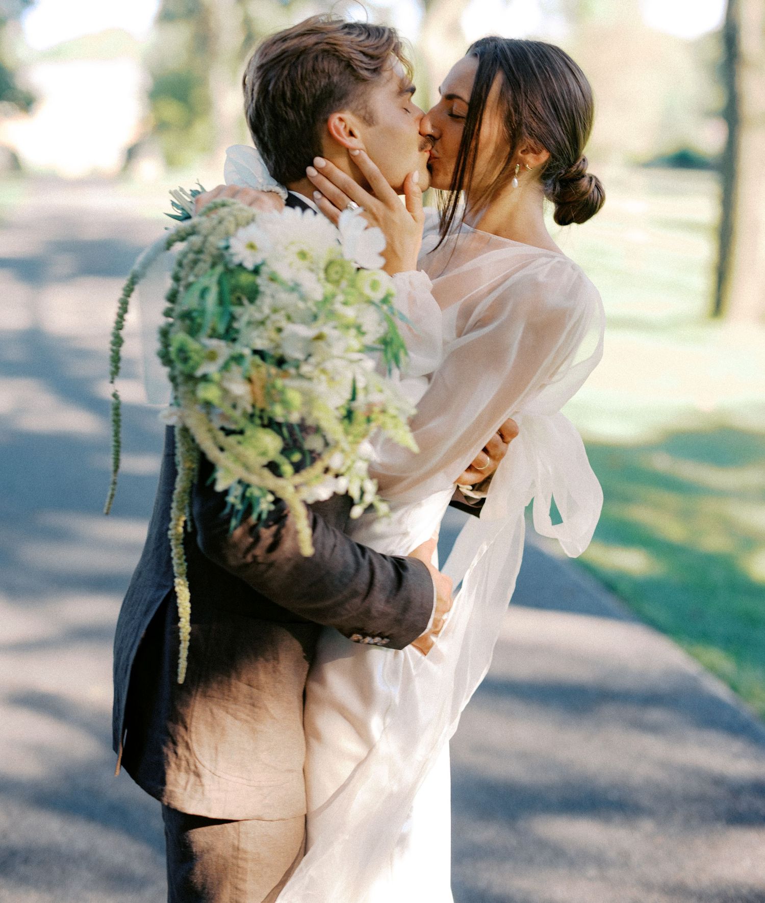 Romantic wedding couple embracing while holding a white floral bouquet outdoors.