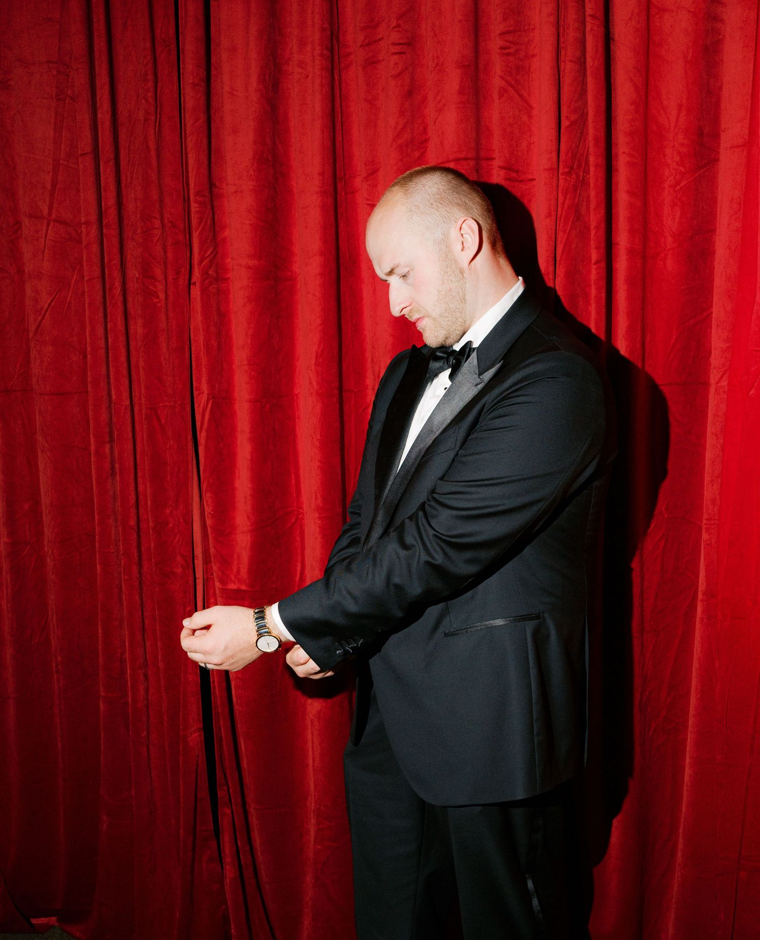 A man in a tuxedo adjusts his cuff links against a red curtain backdrop.