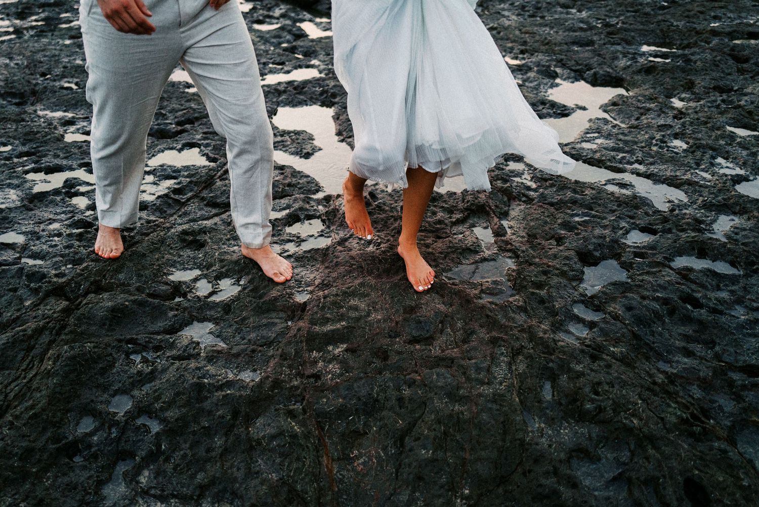 Close-up of feet walking on black volcanic rocks near the ocean's edge.