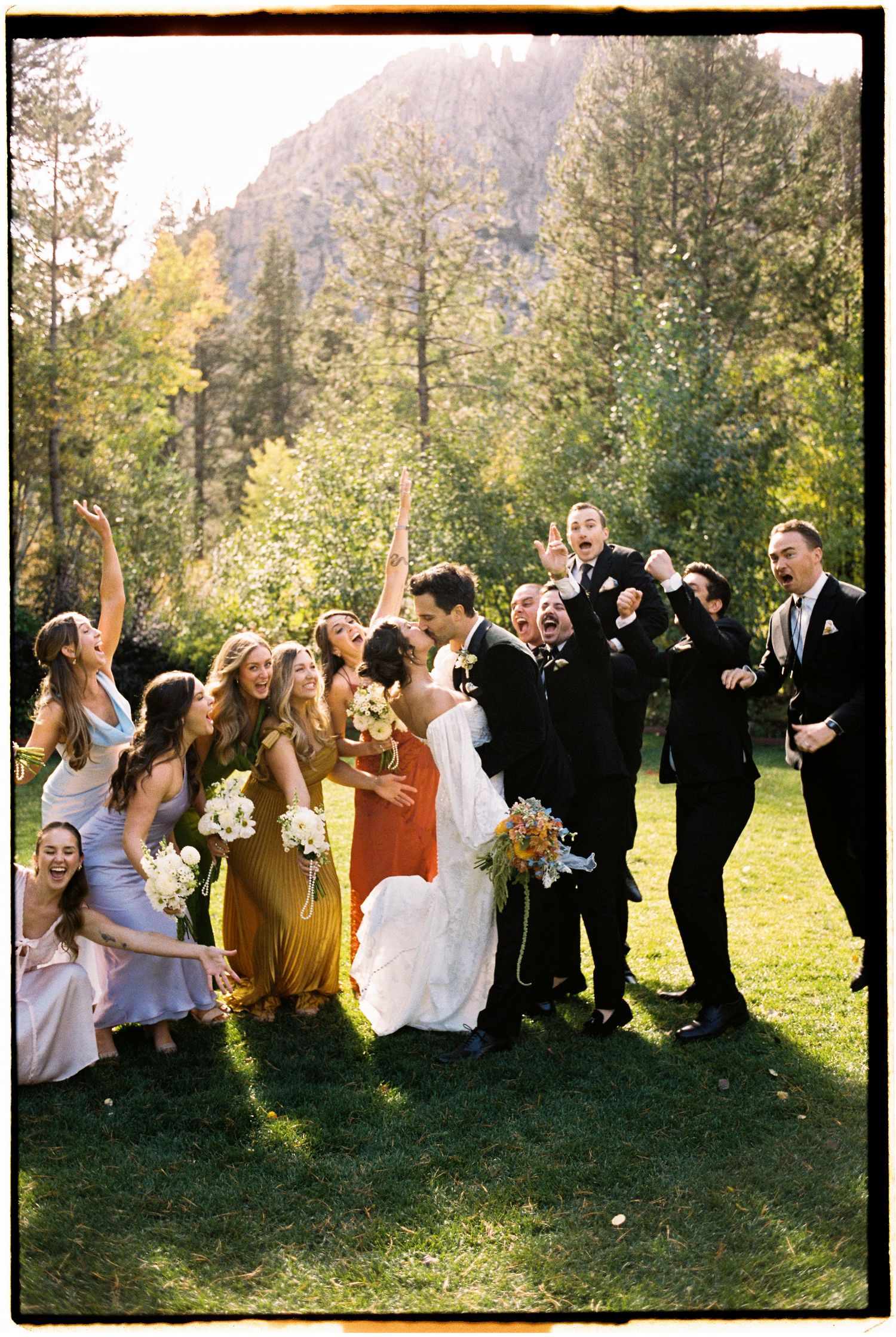 A wedding party poses together on grass with mountains and autumn trees in the background.