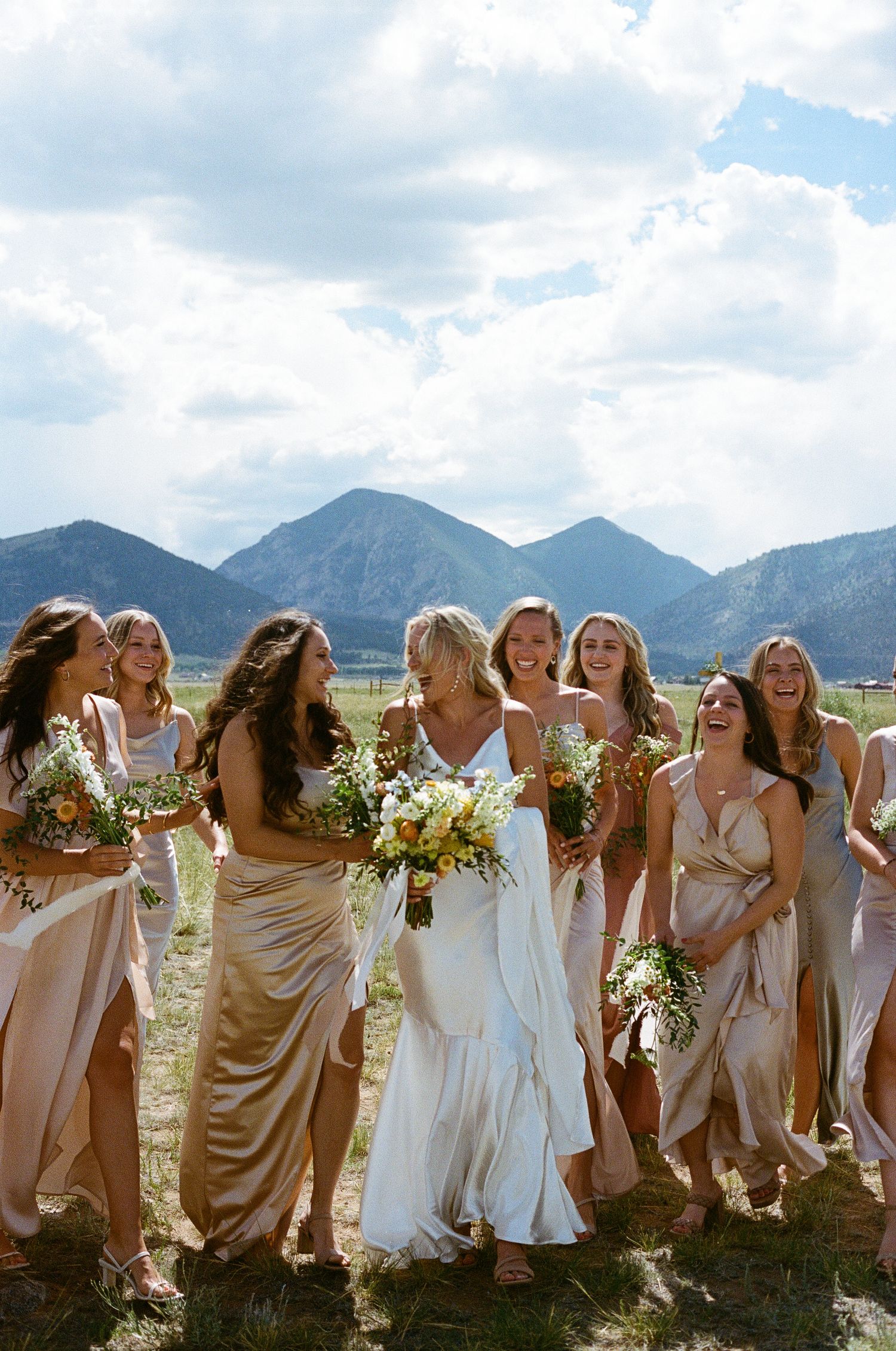 A wedding party in white and beige dresses laughs together against a backdrop of mountain peaks and blue sky.