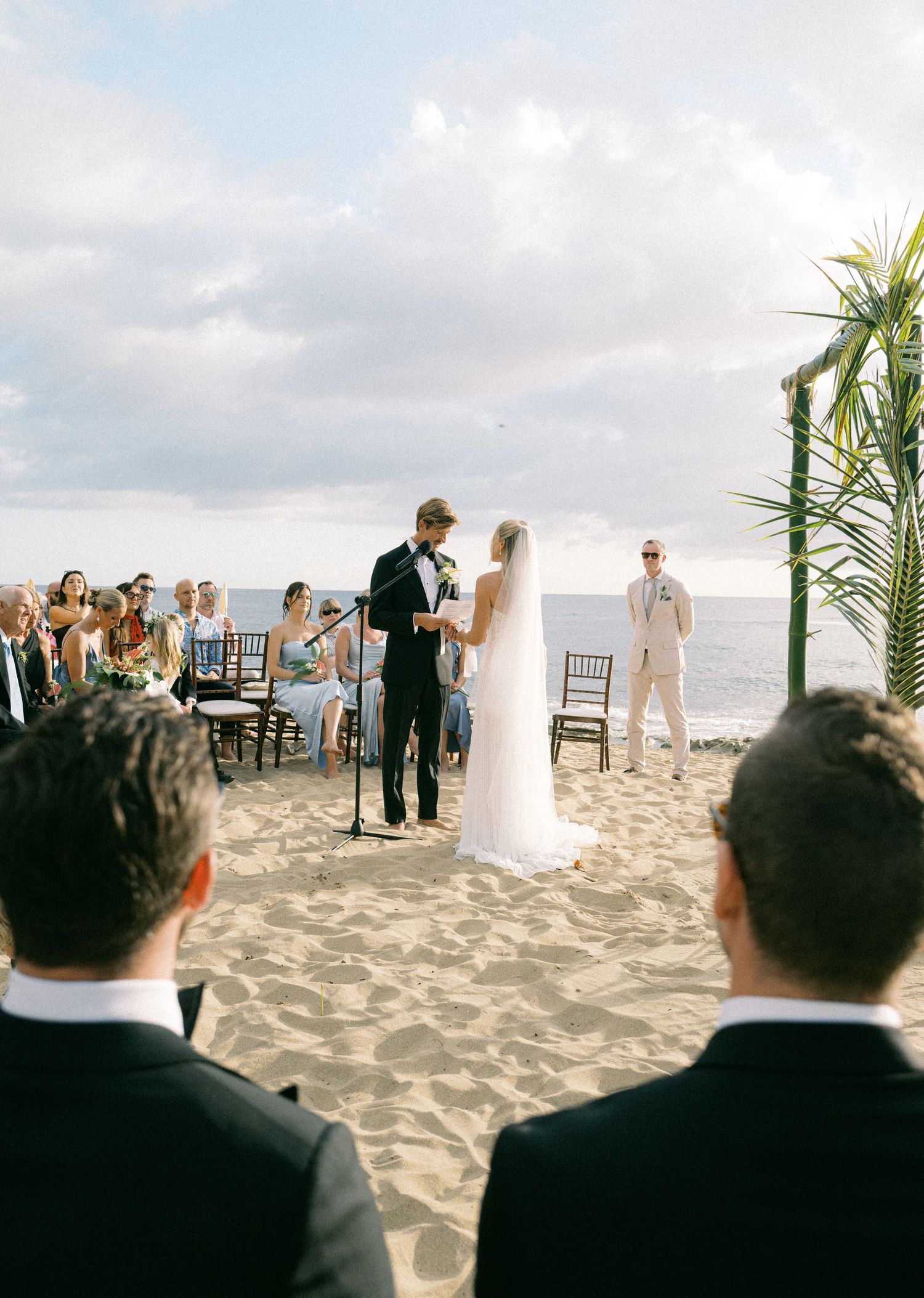 Wedding ceremony taking place on a sandy beach with guests seated in dark chairs and tropical plants as decor.