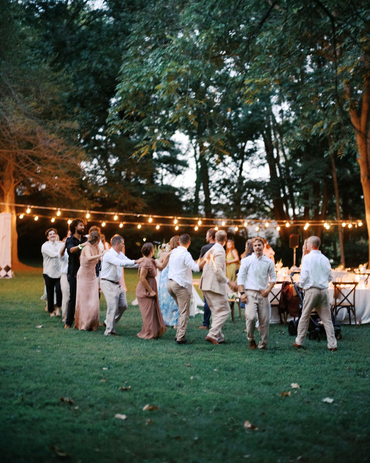 Wedding party dances in a line on a grassy lawn under string lights at dusk during outdoor celebration.
