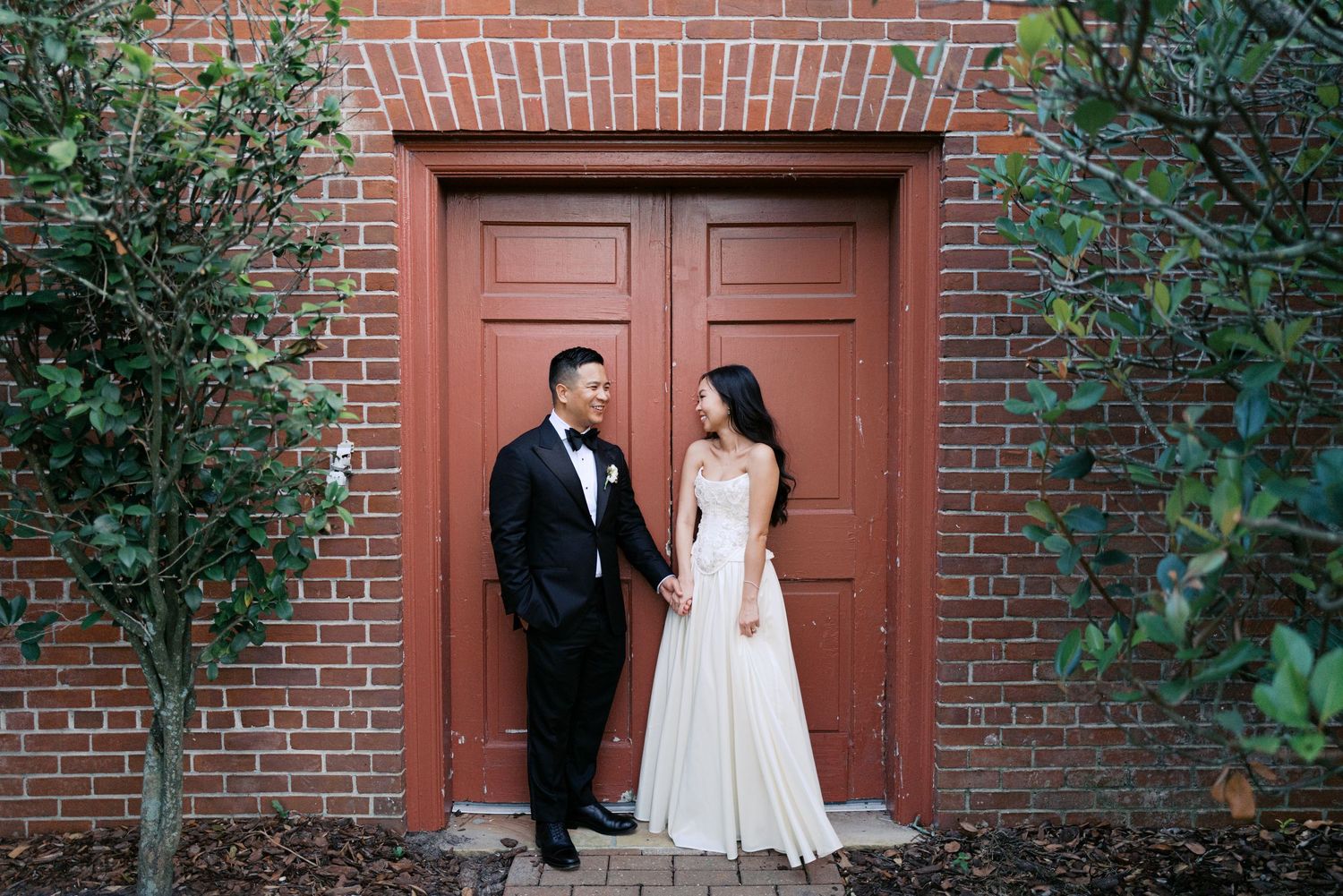 A couple poses elegantly against a rustic brick wall with wooden doors and greenery.