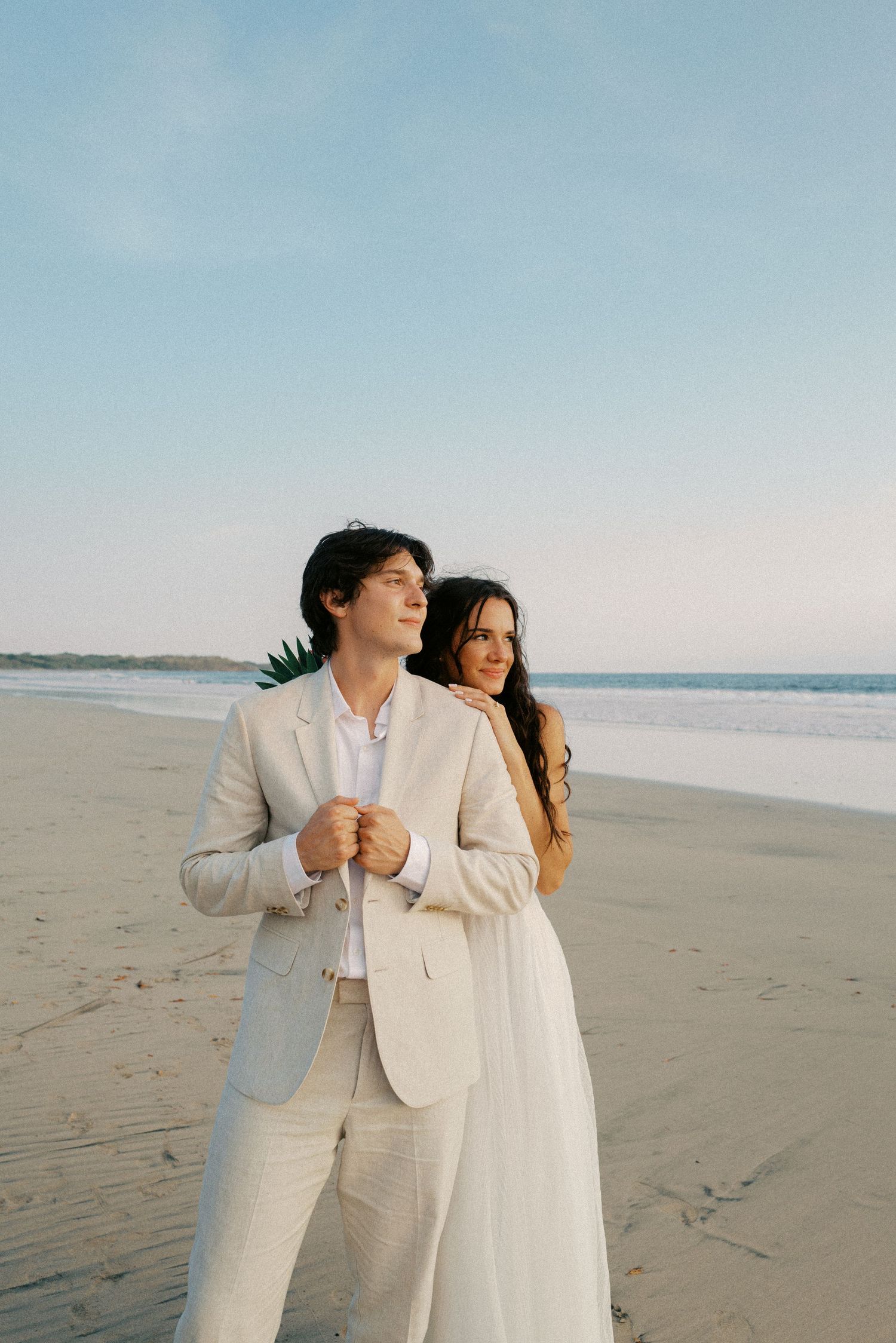 A couple shares an intimate moment on a serene beach with soft blue sky in the background.