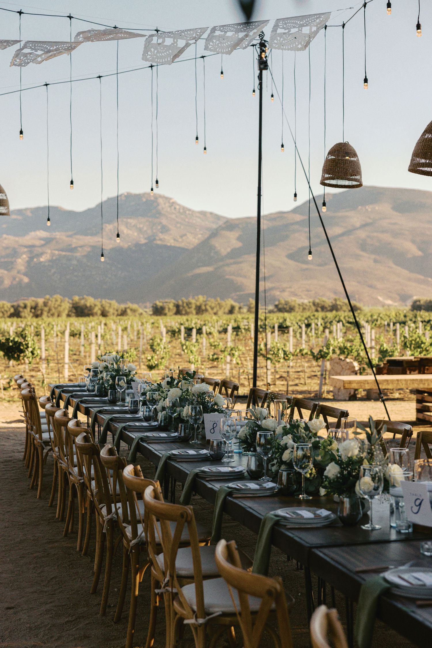 Long rustic dining tables set for an outdoor wedding reception with string lights and mountain views at sunset.
